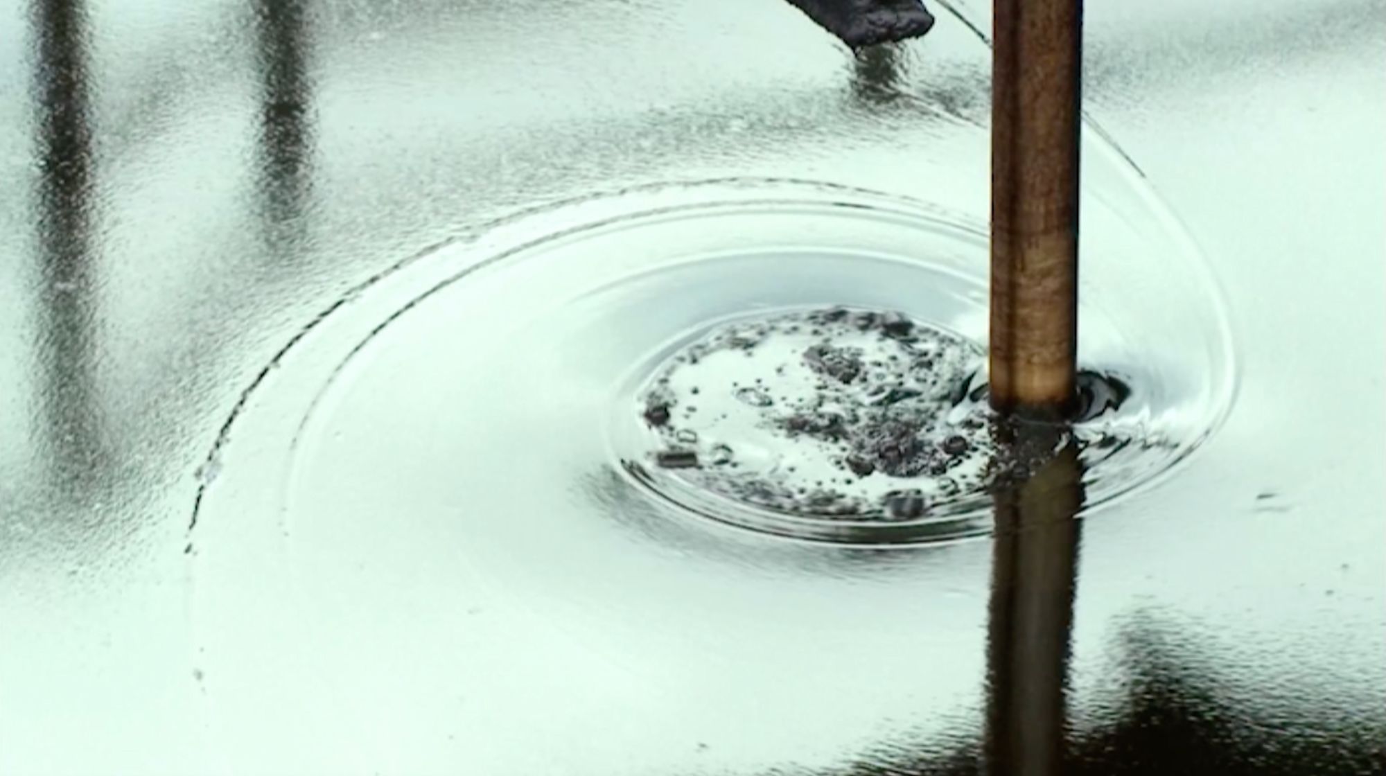 Nahaufnahme: ein Metallstab taucht in gekräuseltes Wasser mit dunklen Ölresten in der Mitte