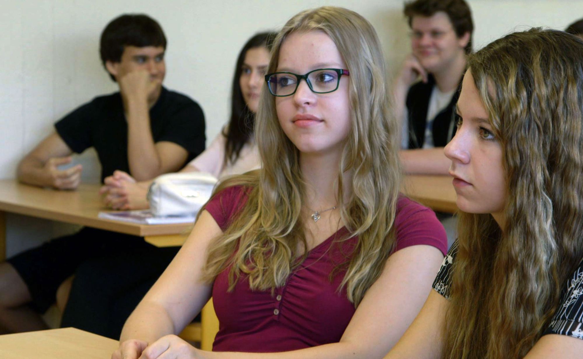 Schülerin mit Brille sitzt aufmerksam im Klassenzimmer, im Hintergrund weitere Jugendliche