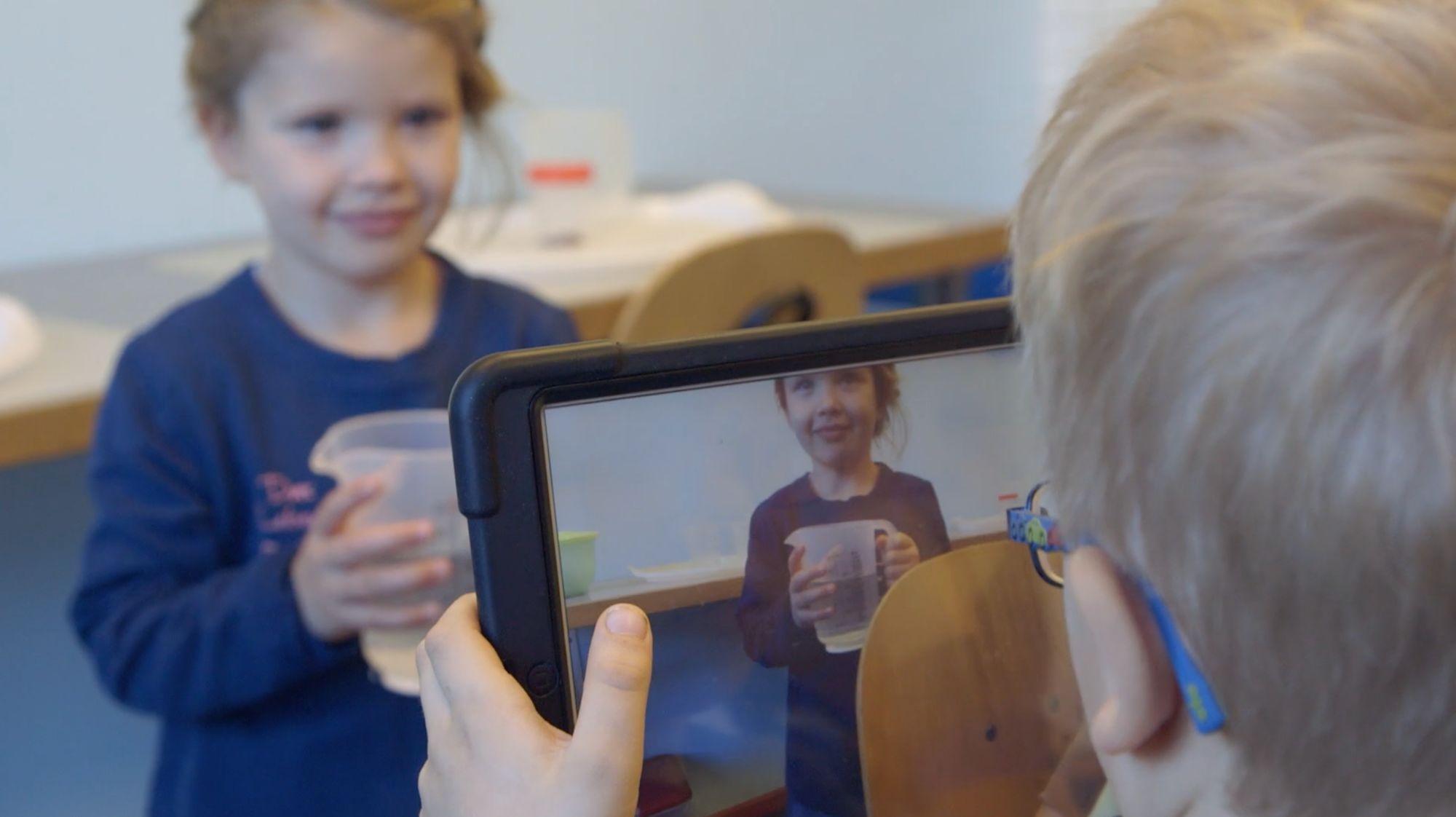 Mädchen mit Becher wird von einem Jungen mit einem Tablet gefilmt, Bild erscheint auf dem Display