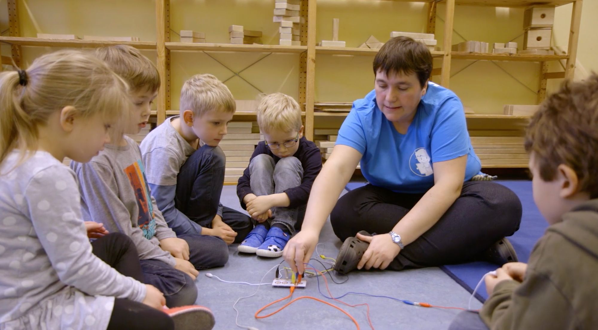 Erzieherin sitzt mit sechs Kindern im Kreis auf dem Boden und experimentiert mit einem Stromkreis-Bausatz