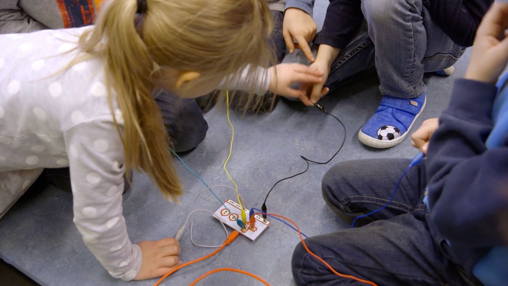 Kinderhände verbinden bunte Krokodilklemmen an einem Experimentierboard auf dem Fußboden