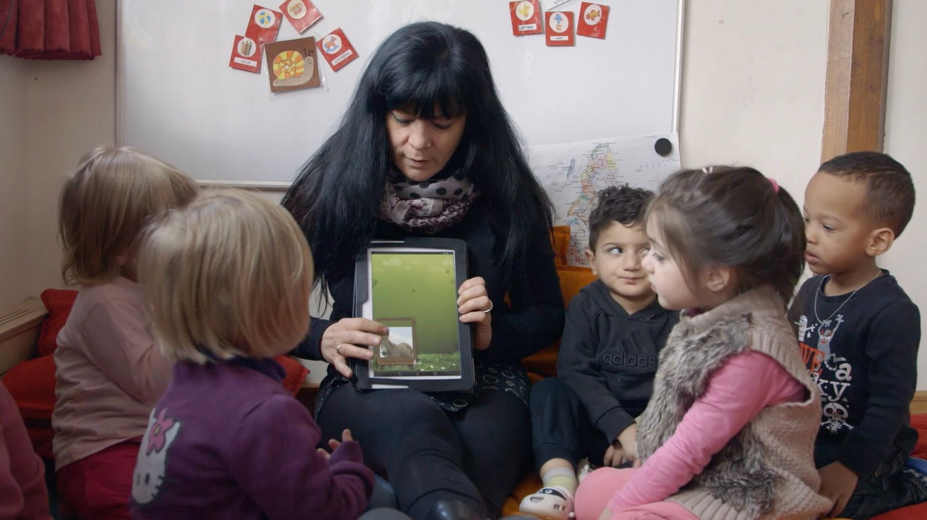 Frau zeigt mehreren kleinen Kindern ein Tablet vor einer Pinnwand mit Namenskärtchen
