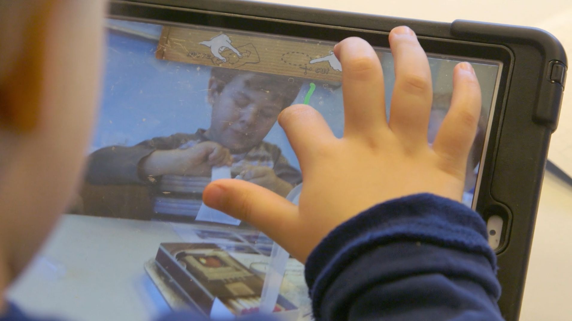 Kinderhand berührt ein Tablet, auf dessen Display ein Kind beim Basteln zu sehen ist