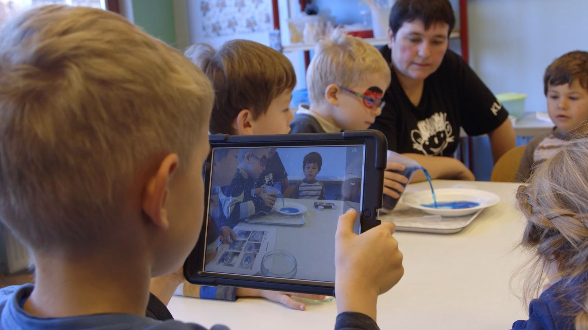 Junge hält ein Tablet mit einer Videoaufnahme von Kindern, die blaue Farbe in eine Schale gießen
