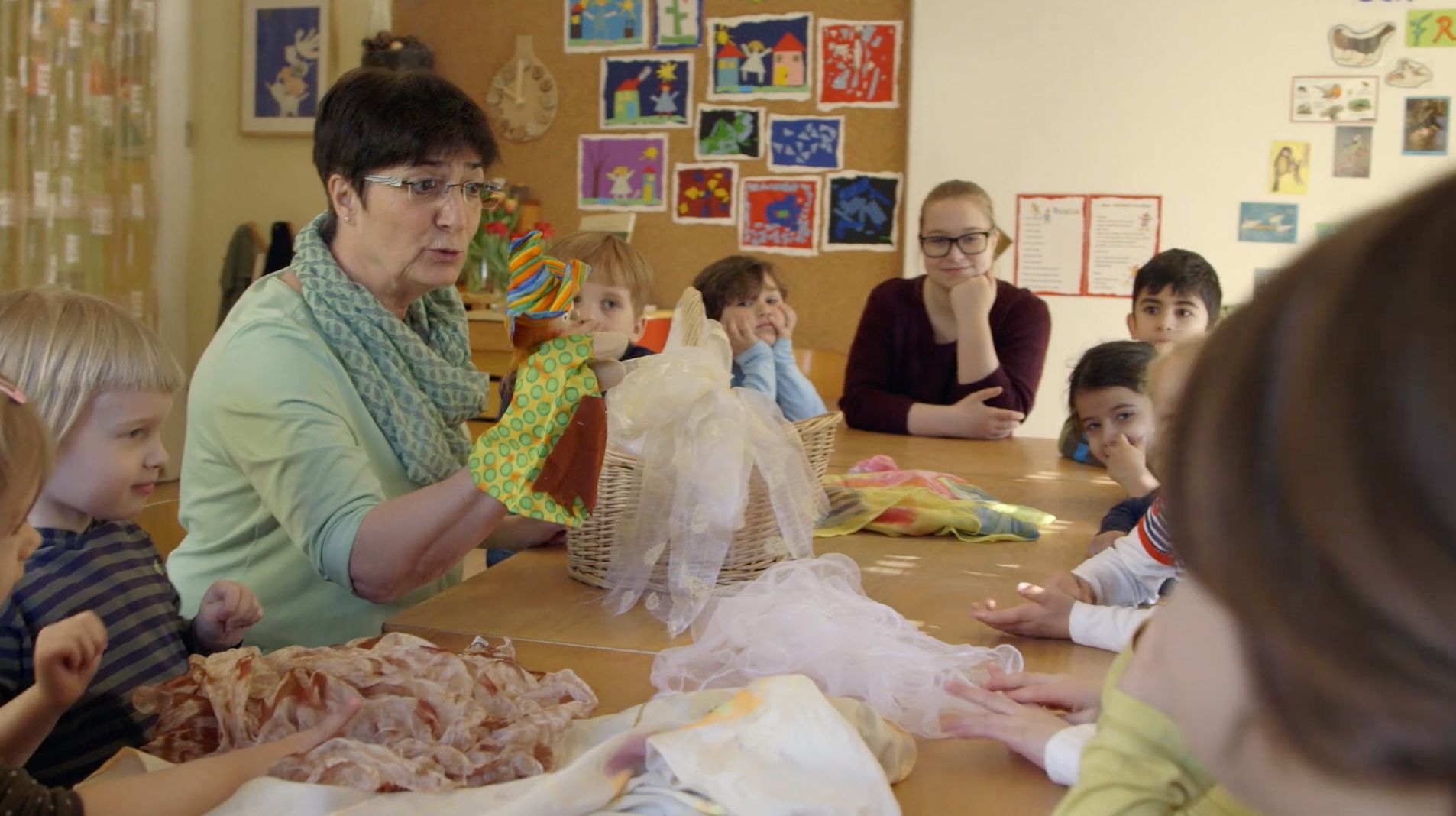 Frau mit Handpuppe erzählt einer Kindergruppe am Tisch, durchsichtige Tücher liegen vor ihnen