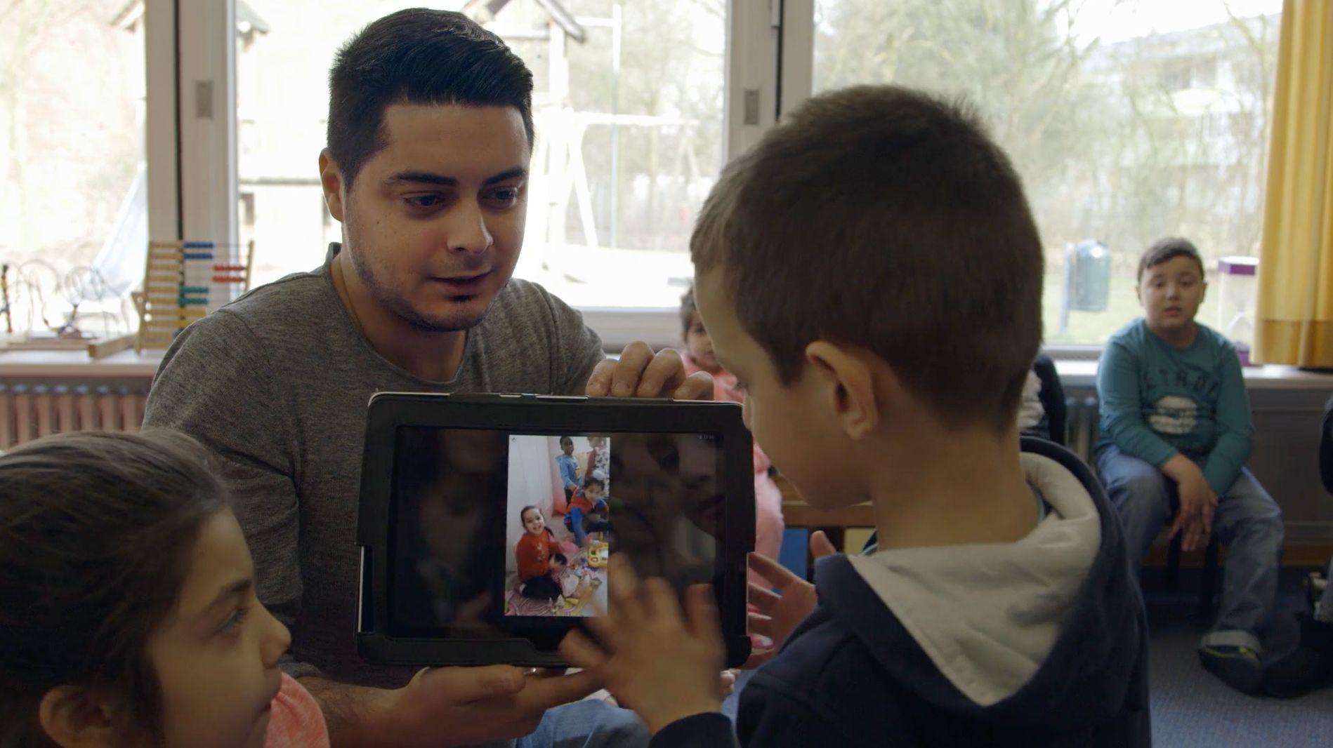 Mann zeigt zwei Kindern Fotos auf einem Tablet, weitere Kinder sitzen im Hintergrund