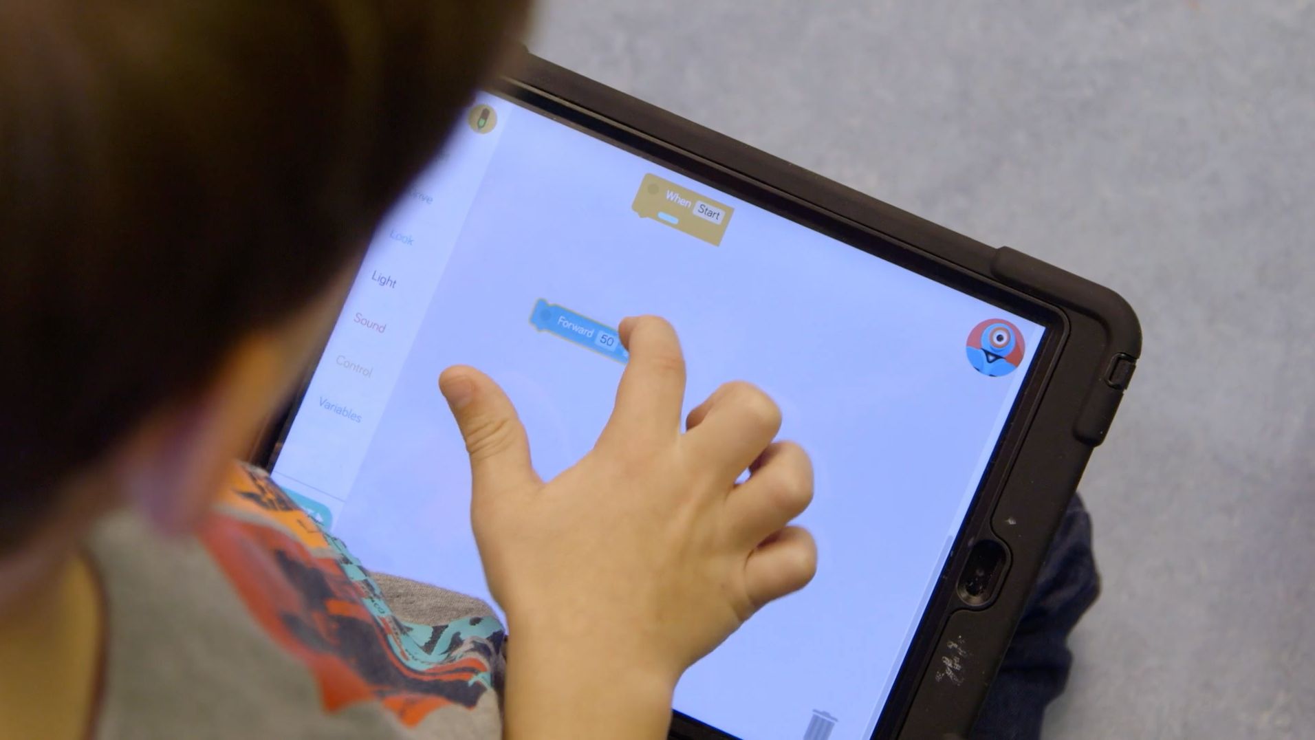 Kinderhand berührt ein Tablet mit einer Programmier-App, Codeblöcke wie „Forward 50“ sind zu sehen