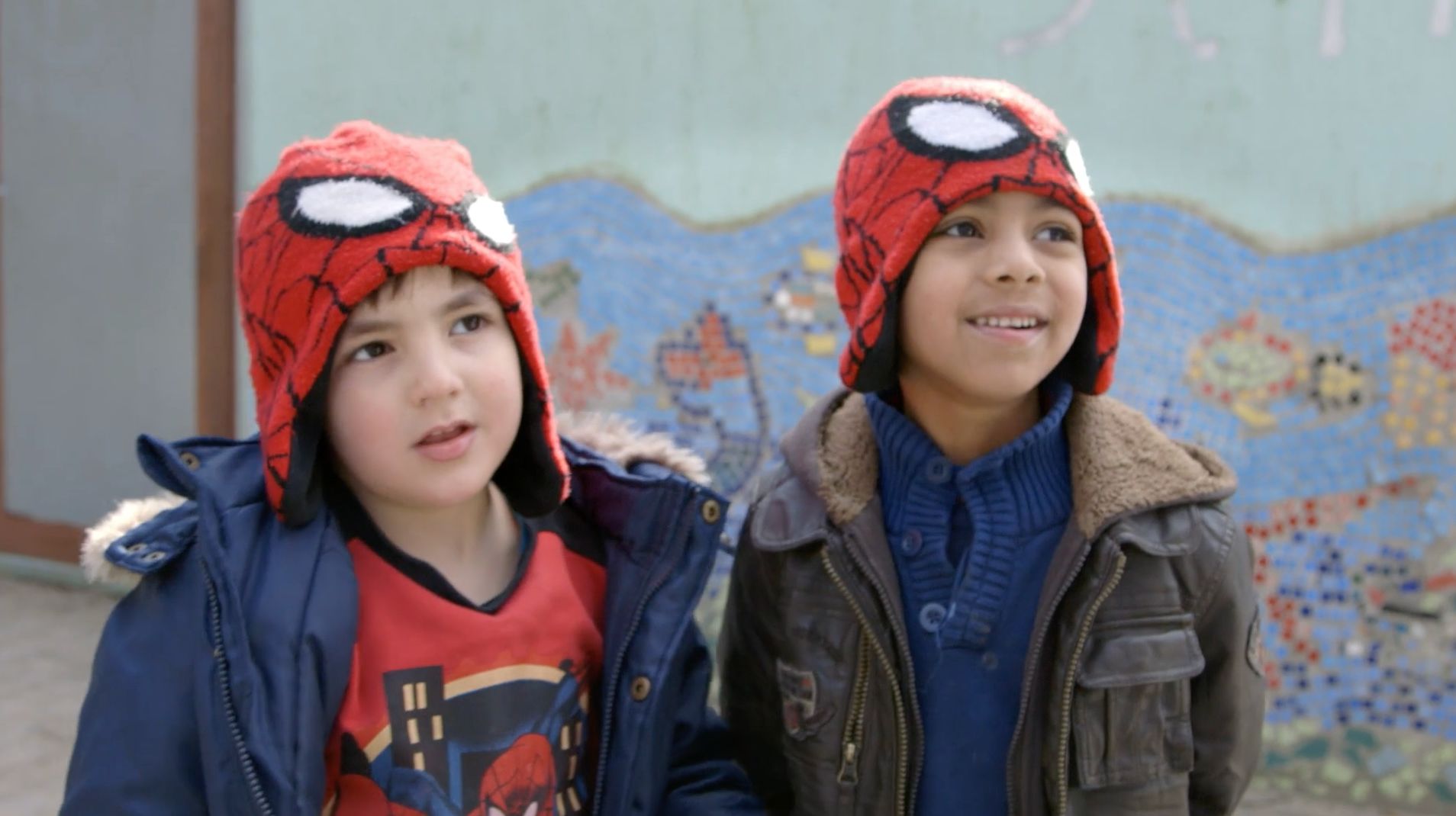 Zwei Jungen mit roten Spiderman-Mützen stehen vor einer bunten Mosaikwand im Freien