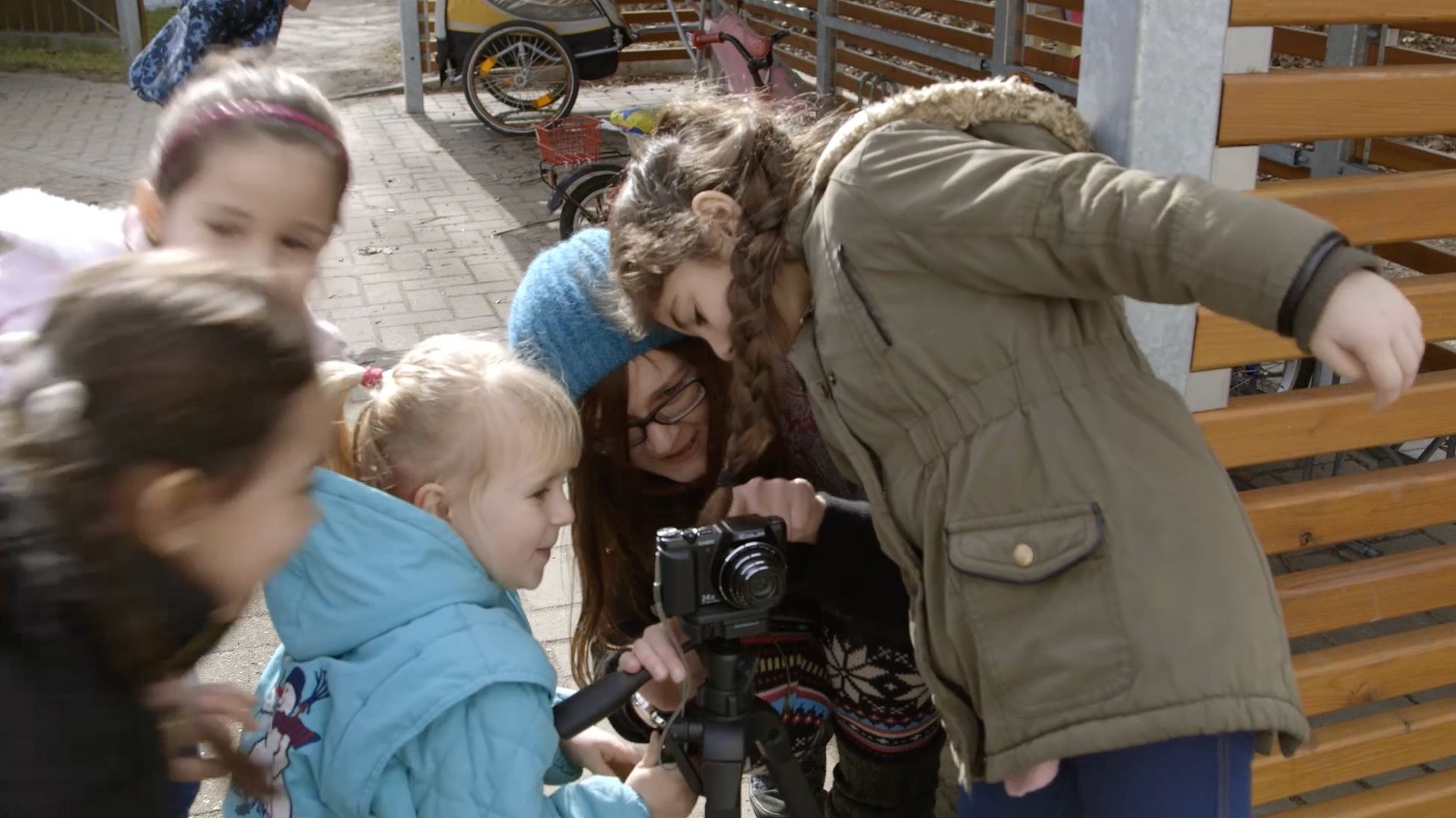 Gruppe von Kindern und eine Frau betrachten gemeinsam Aufnahmen auf einer Kamera im Freien