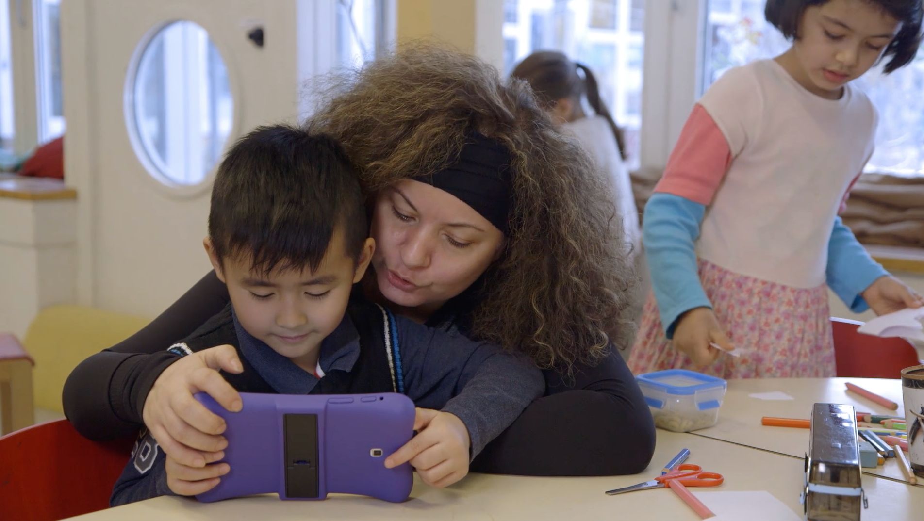 Erzieherin umarmt einen Jungen, gemeinsam schauen sie auf ein lilafarbenes Tablet
