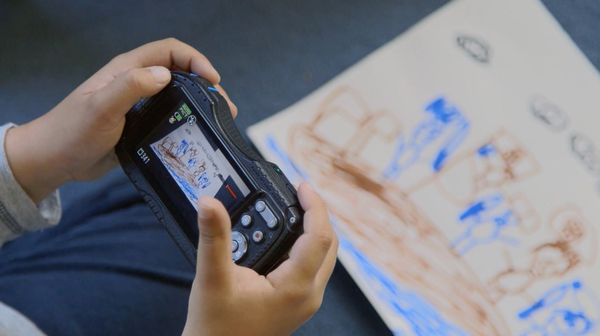 Hände halten eine Digitalkamera, auf deren Display eine bunte Kinderzeichnung zu sehen ist