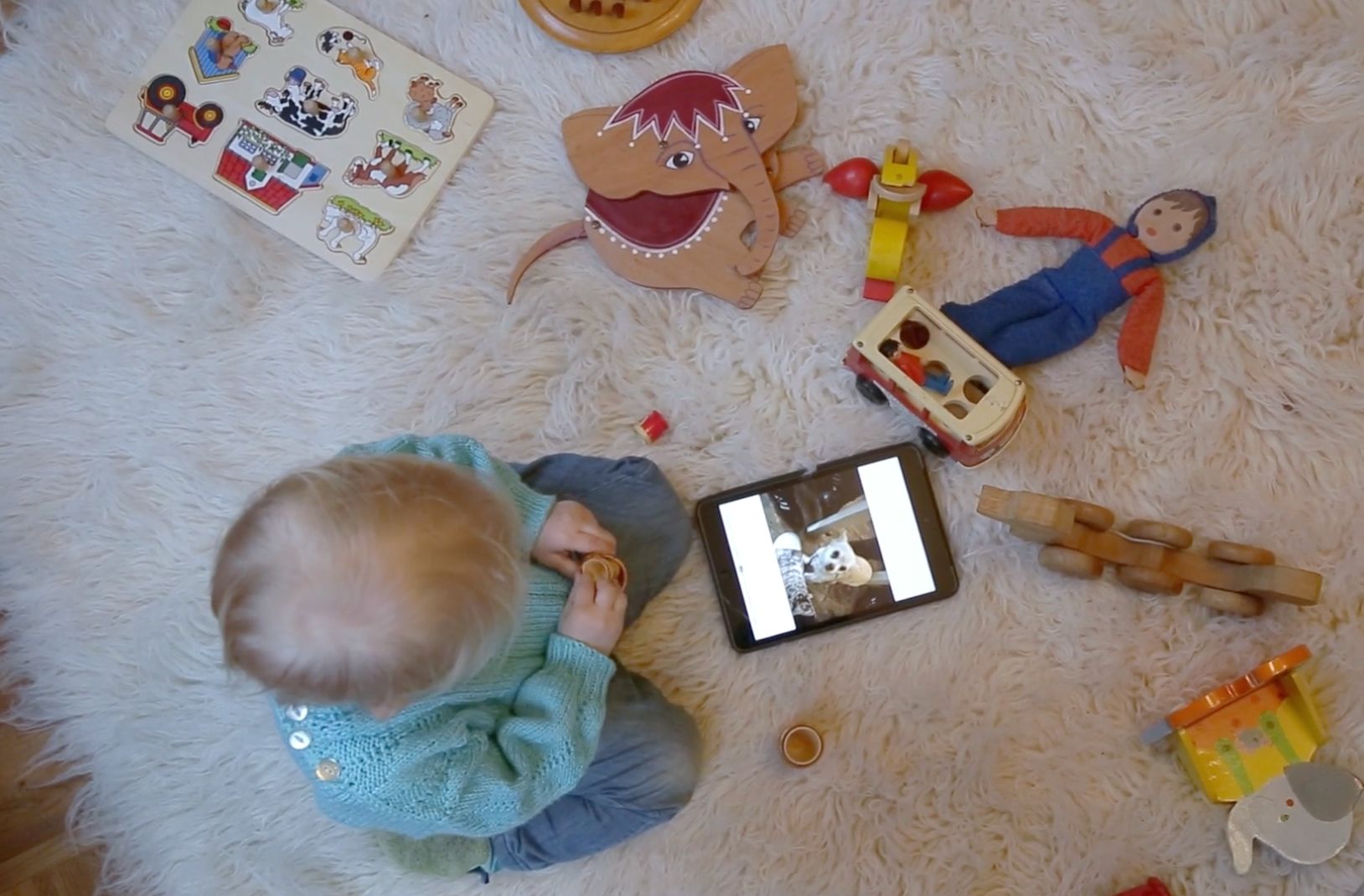 Kleinkind sitzt zwischen Holzspielzeug auf einem Fell und schaut auf ein Tablet