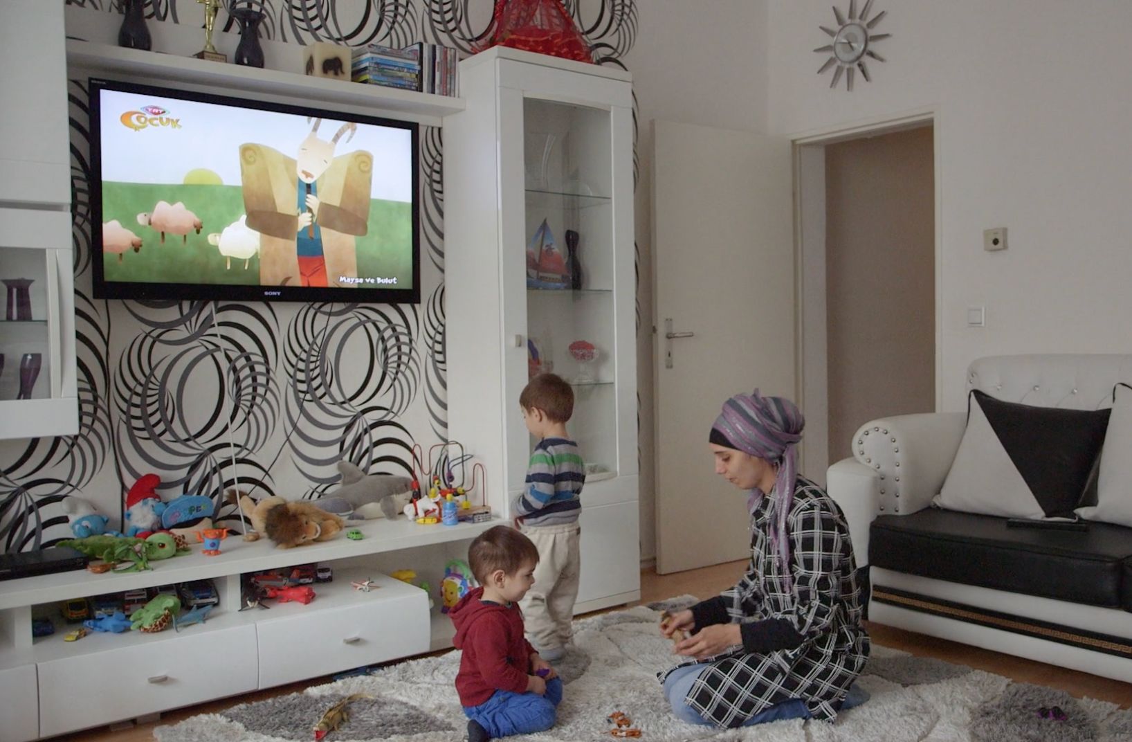 Zwei Jungen und eine Frau mit Kopftuch sitzen im Wohnzimmer vor einem laufenden Zeichentrickfilm