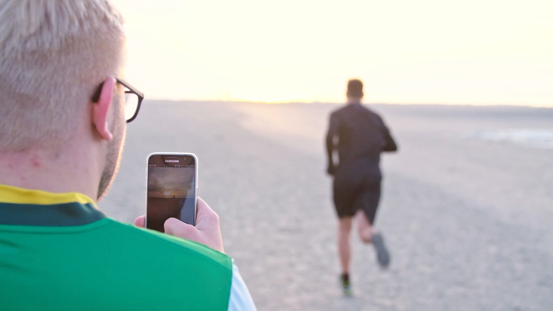Mann fotografiert mit dem Smartphone einen Läufer am Strand bei Sonnenuntergang