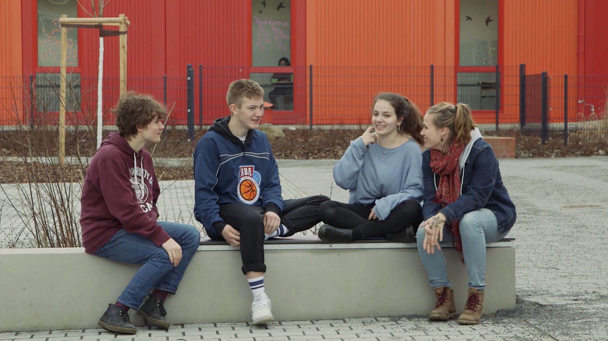 Vier Jugendliche sitzen im Gespräch auf einer Bank vor einem roten Schulgebäude