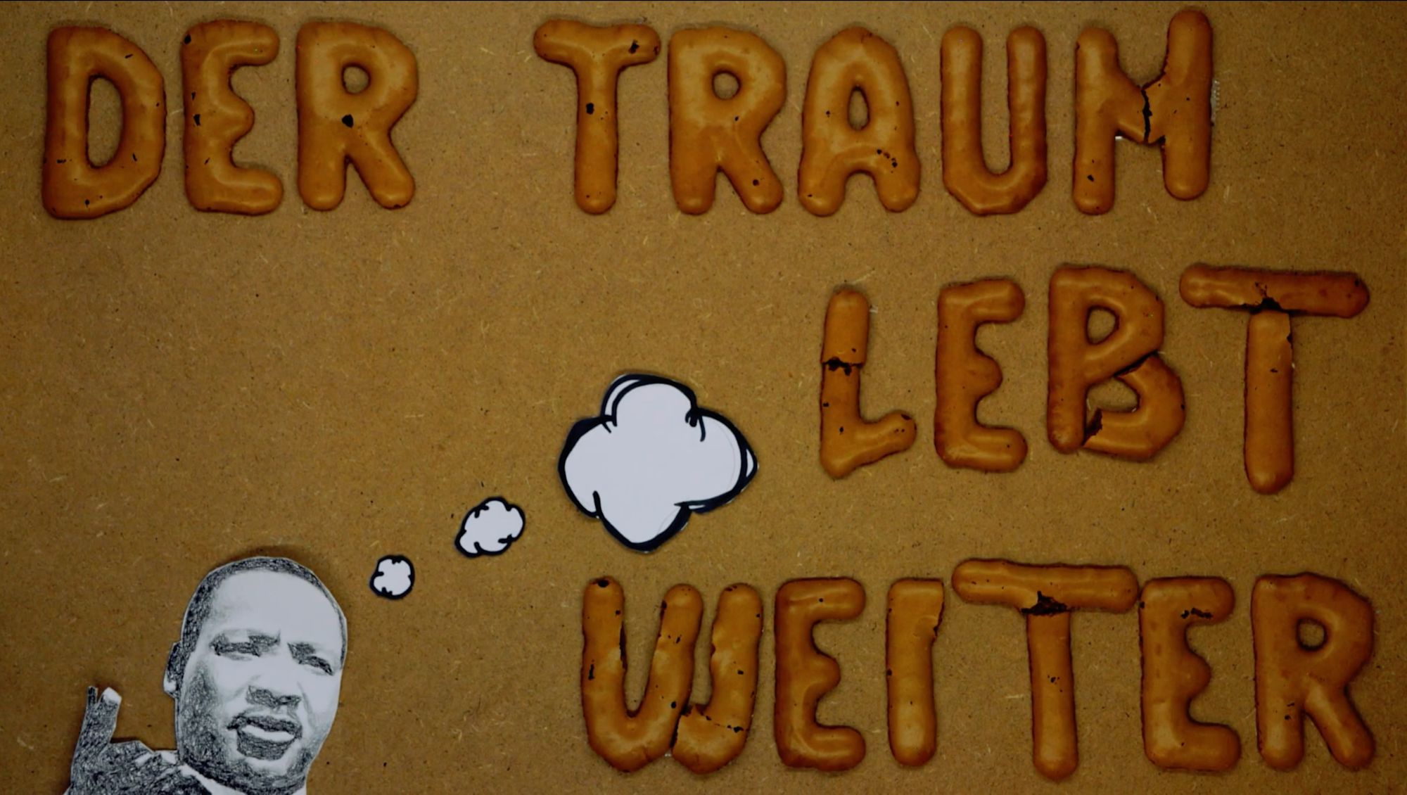 Illustrierte Szene: Schriftzug „Der Traum lebt weiter“ aus Keksbuchstaben mit Porträtzeichnung