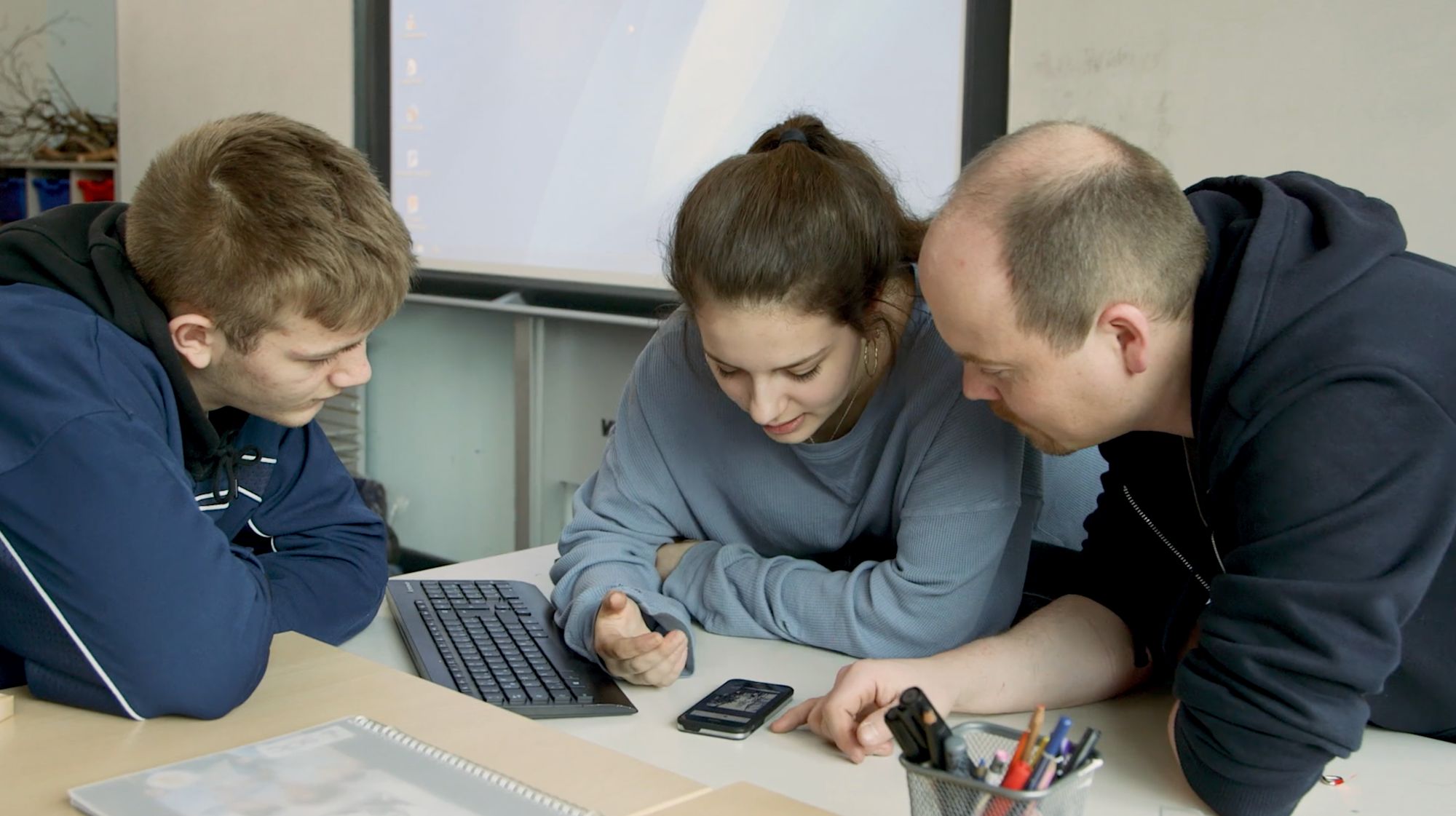 Drei Personen schauen gemeinsam auf einen Laptop und ein Smartphone auf dem Tisch