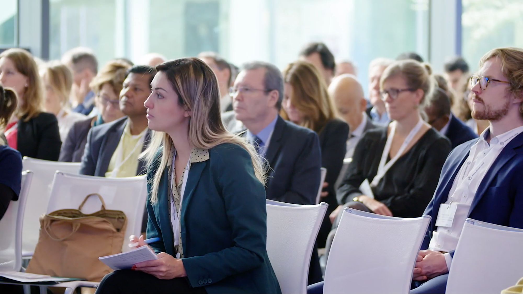 Zuhörer bei einer Konferenz, im Vordergrund eine Frau mit Notizblock