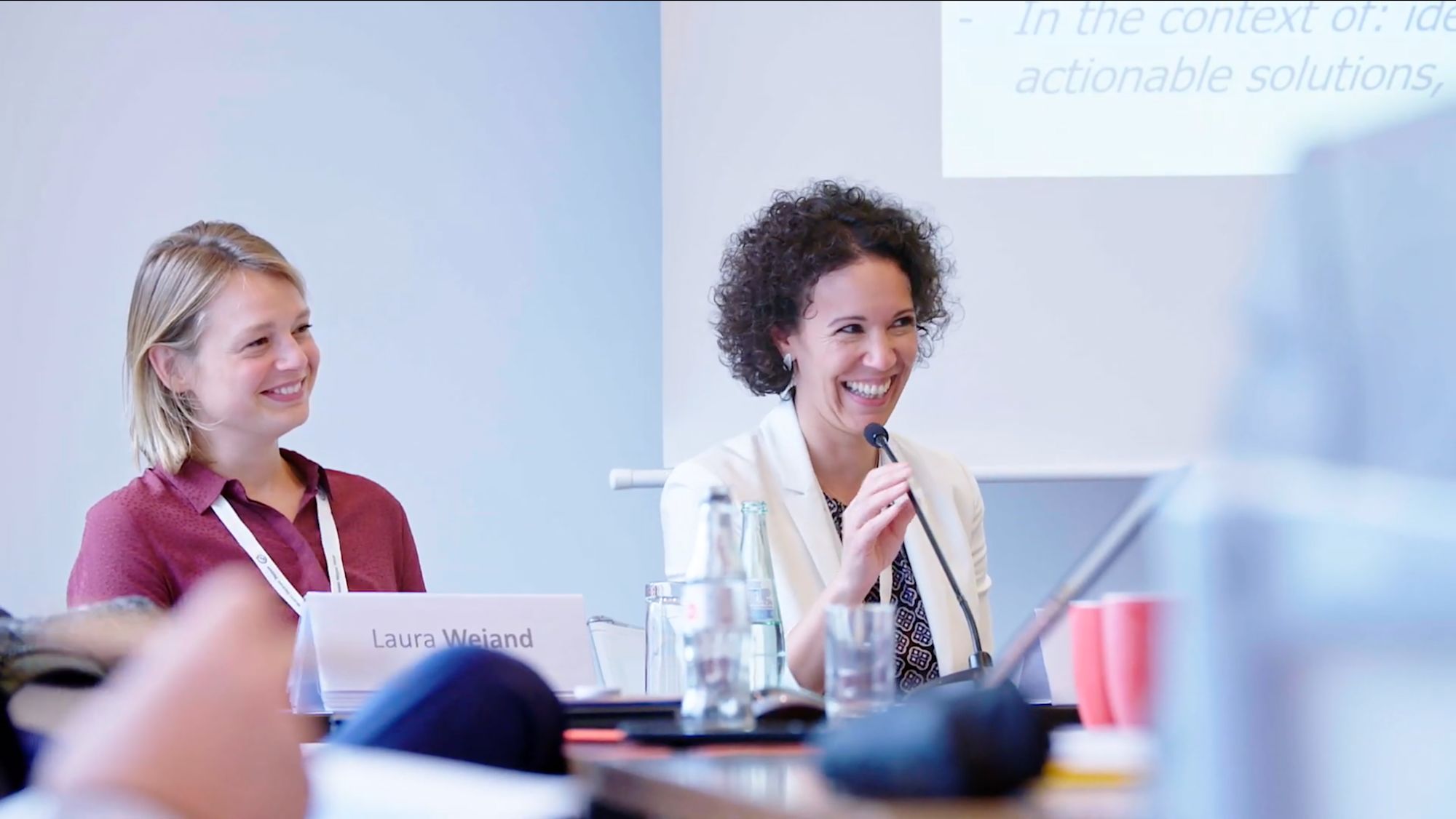 Zwei Frauen auf einem Podium, eine spricht lächelnd ins Mikrofon, Namensschild „Laura Wejand“