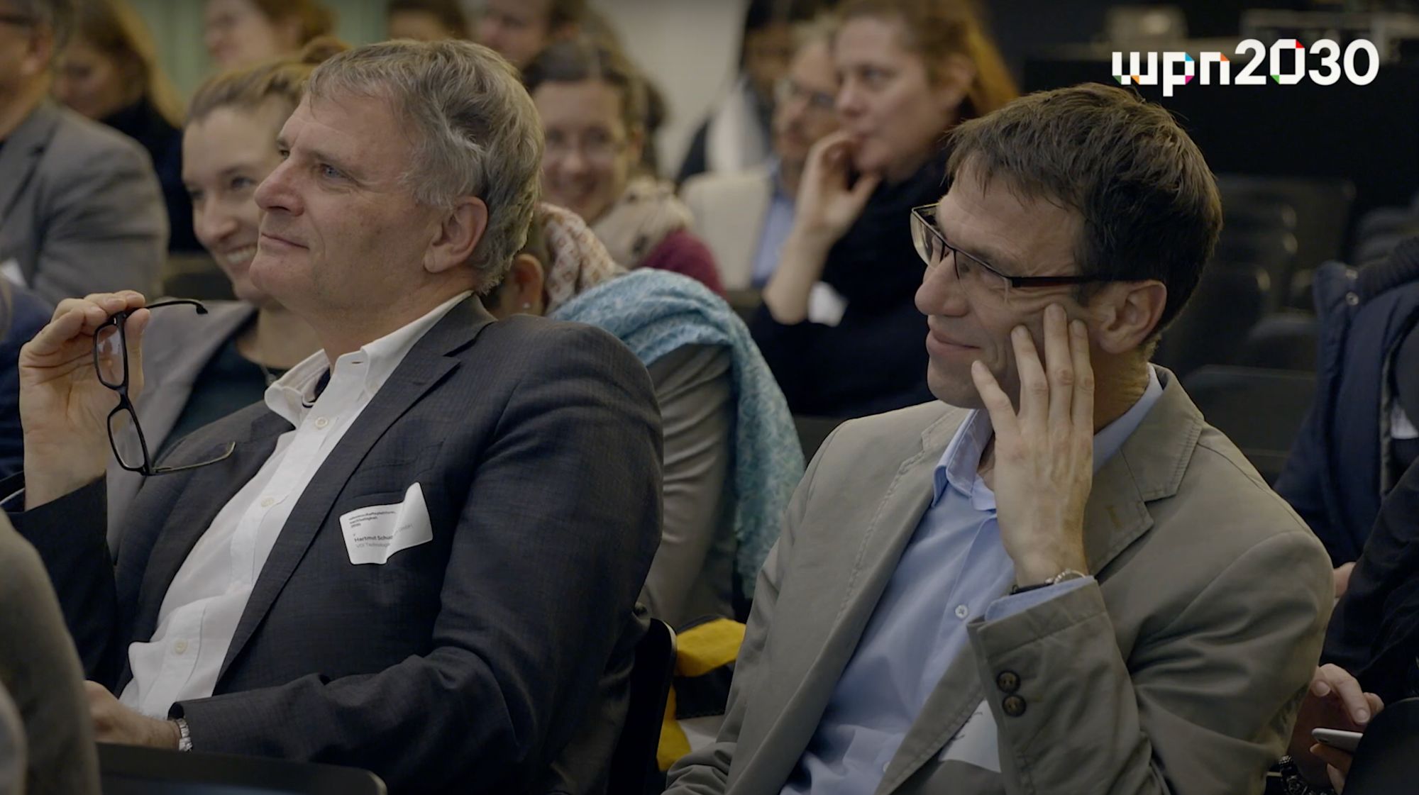 Zwei Männer im Publikum einer Konferenz, einer hält seine Brille in der Hand