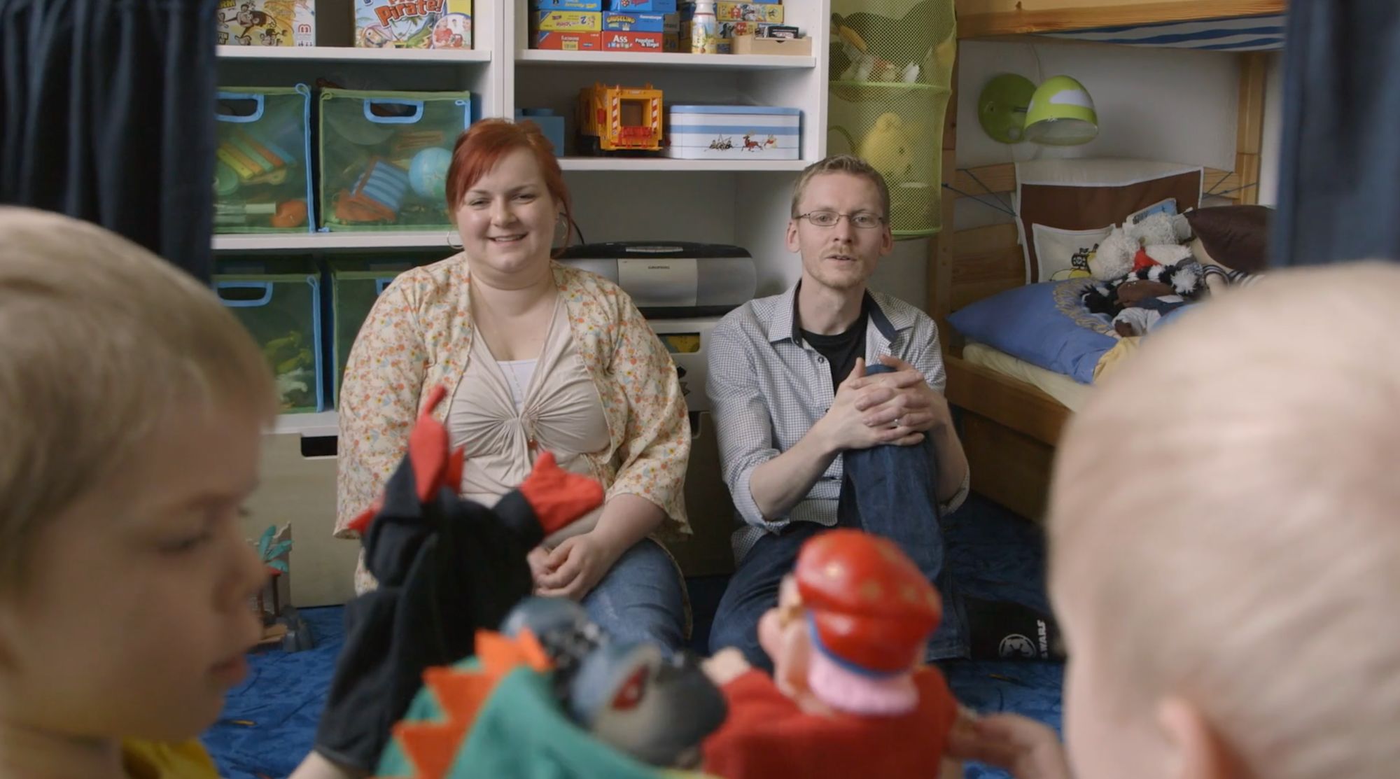 Eltern sitzen im Kinderzimmer, im Vordergrund spielen Kinder mit Handpuppen