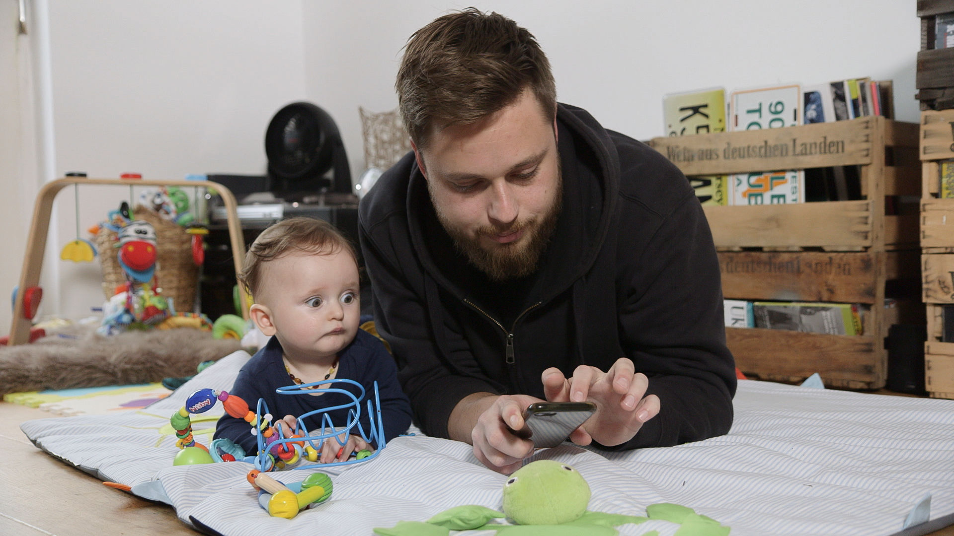 Vater liegt mit Baby auf einer Krabbeldecke und schaut aufs Smartphone