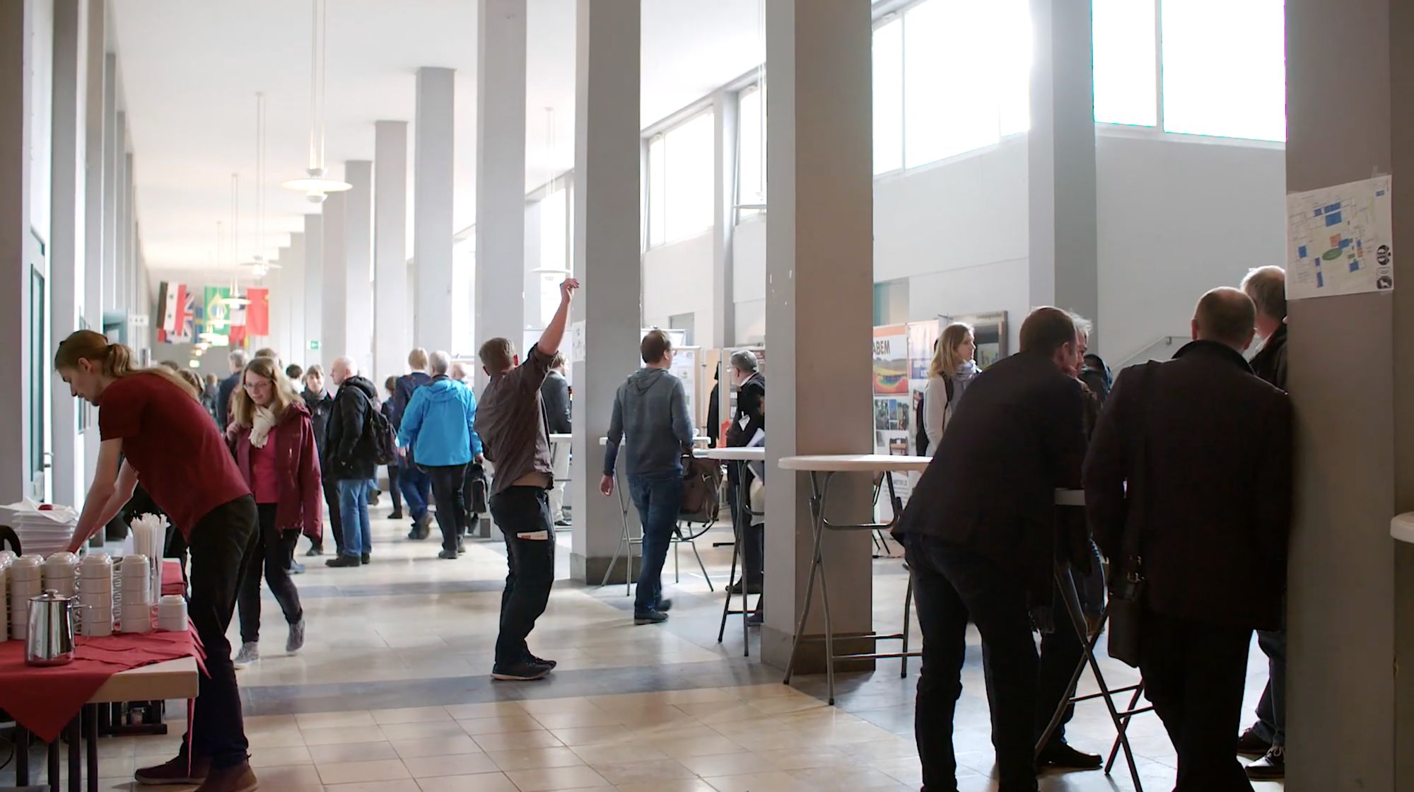 Publikum wandelt durch eine lichte Säulenhalle mit Infoständen und Flaggen bei einer Tagung