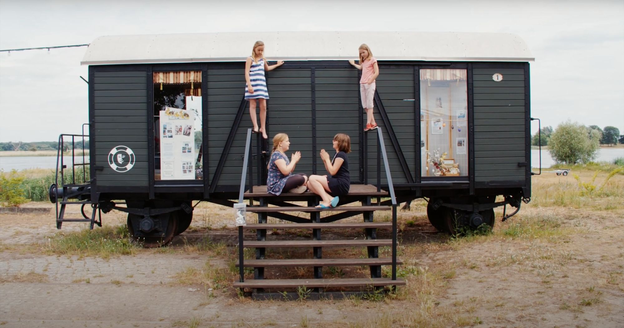 Alter Eisenbahnwaggon am Wasser, zwei Mädchen klettern aufs Dach, zwei Frauen gestikulieren auf der Treppe