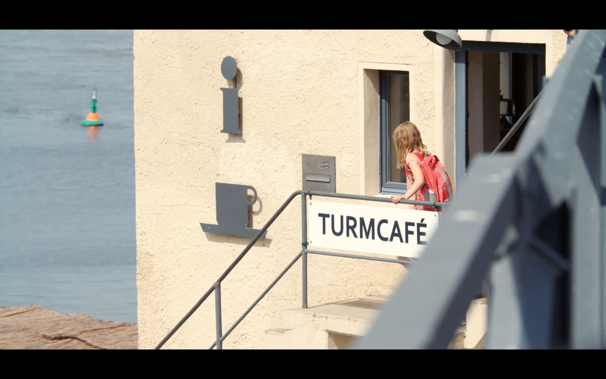 Mädchen mit rosa Rucksack blickt durchs Fenster neben dem Schild „Turmcafé“ am Wasser