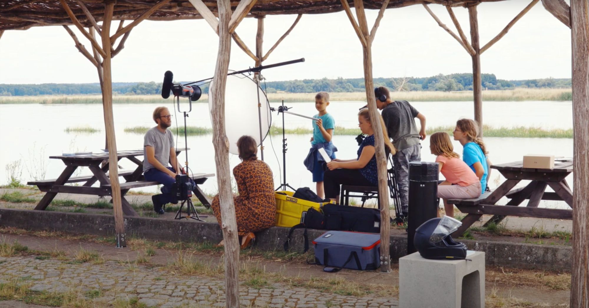 Filmteam mit Kamera und Lichtequipment interviewt Personen unter einem Holzpavillon am See