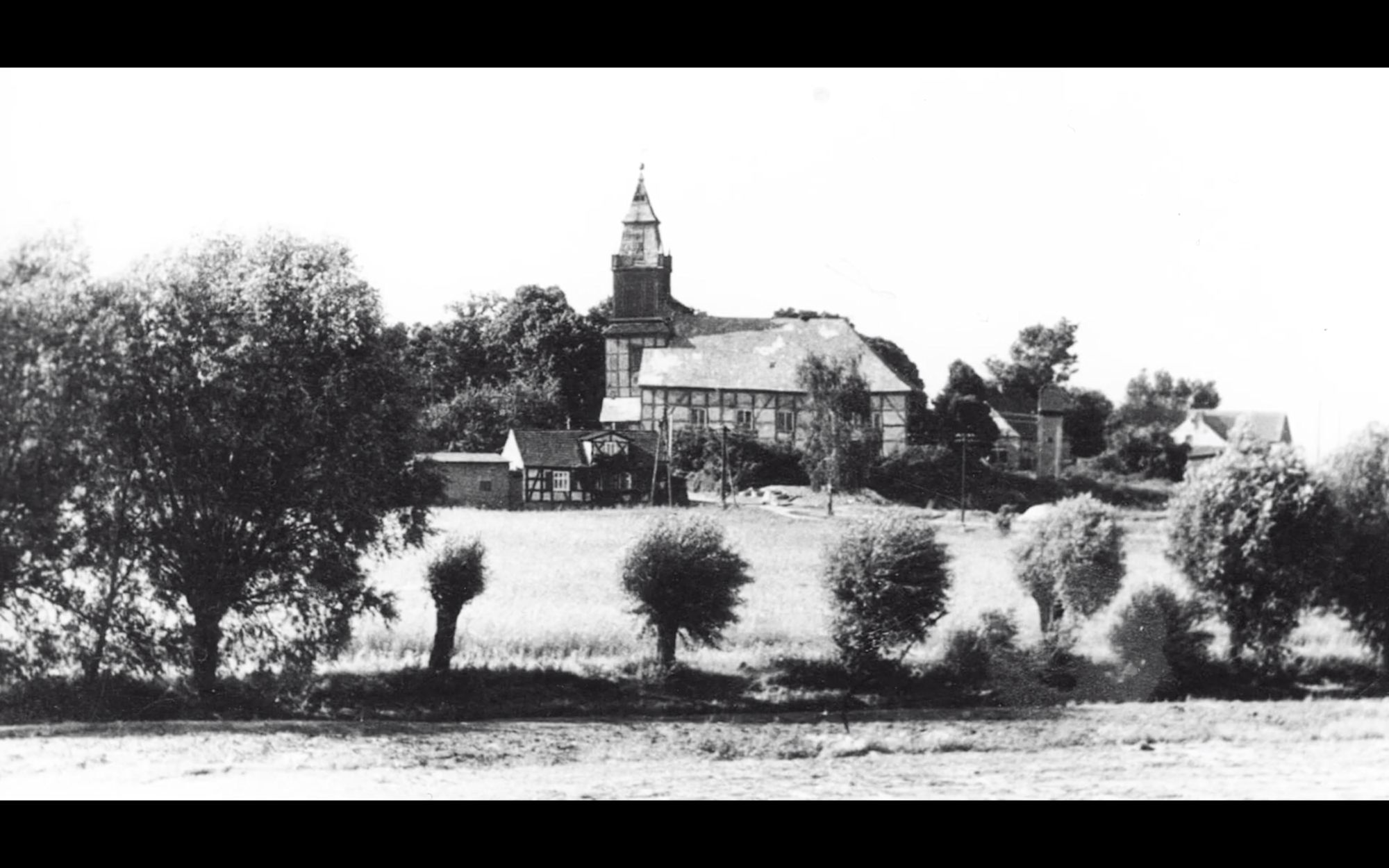 Historische Schwarz-Weiß-Aufnahme einer Dorfkirche mit Fachwerkhäusern und Kopfweiden