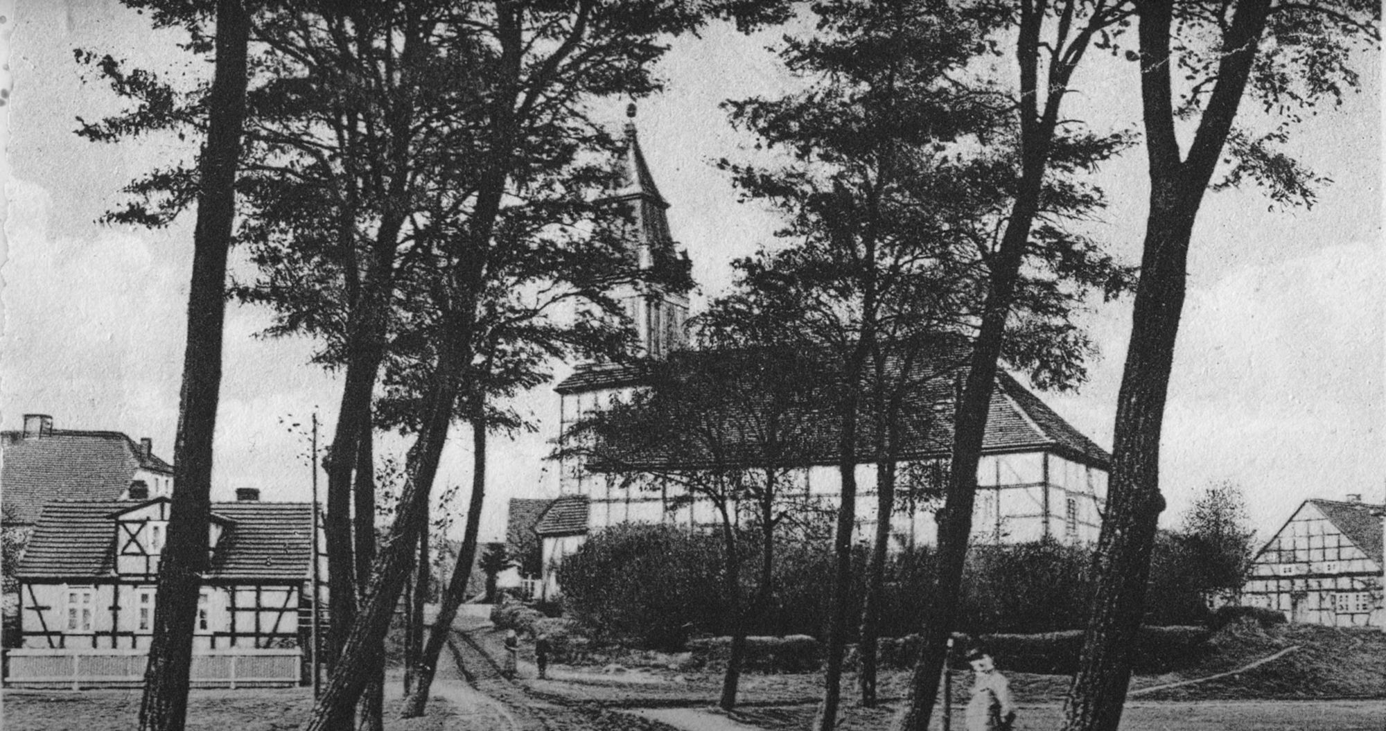 Historische Aufnahme eines Kirchturms zwischen hohen Bäumen an einem Weg