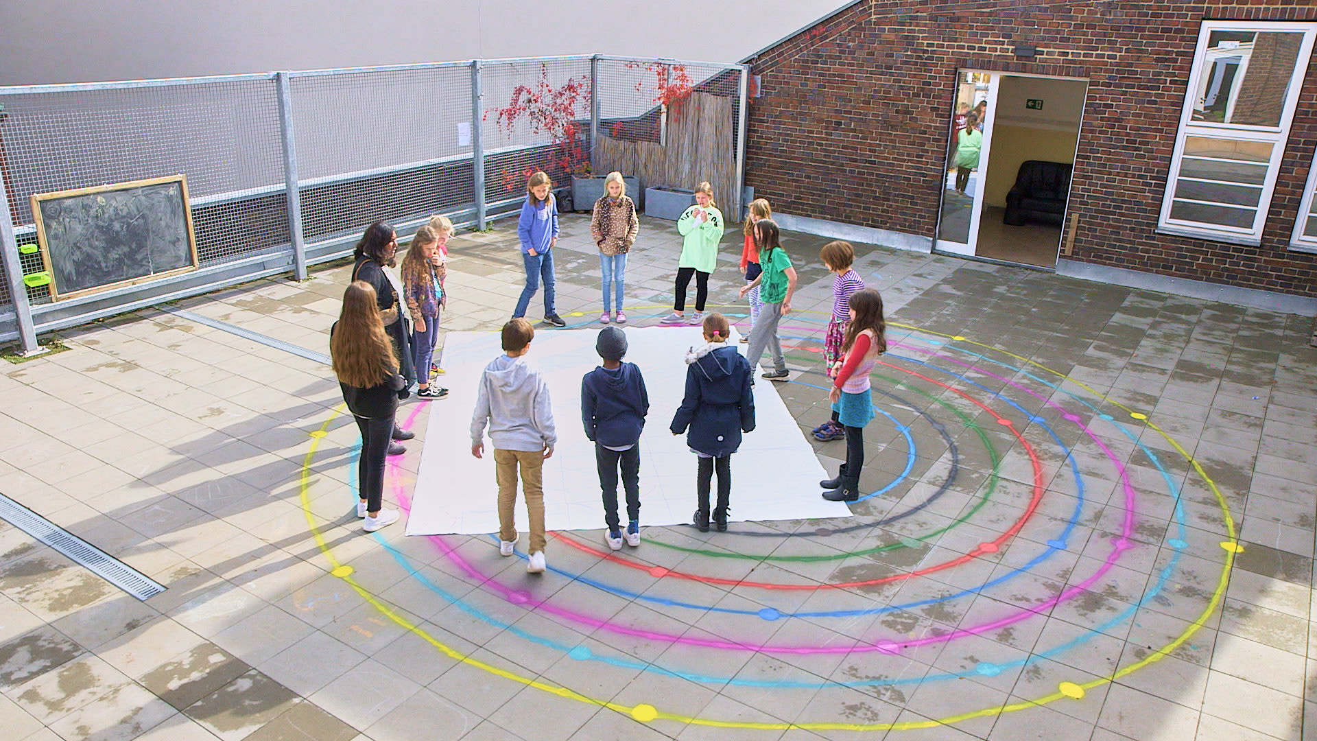 Luftaufnahme: Kinder stehen auf einem Schulhof neben bunten, konzentrischen Kreisen aus Kreide