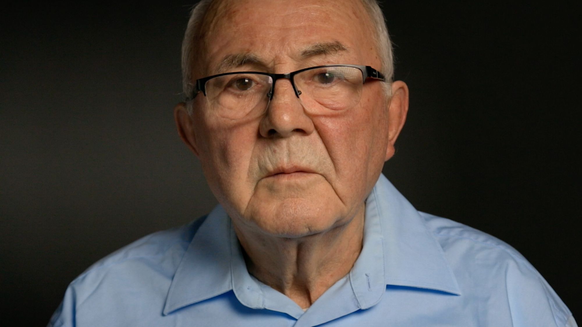 Nahaufnahme eines älteren Mannes mit Brille und hellblauem Hemd vor dunklem Hintergrund