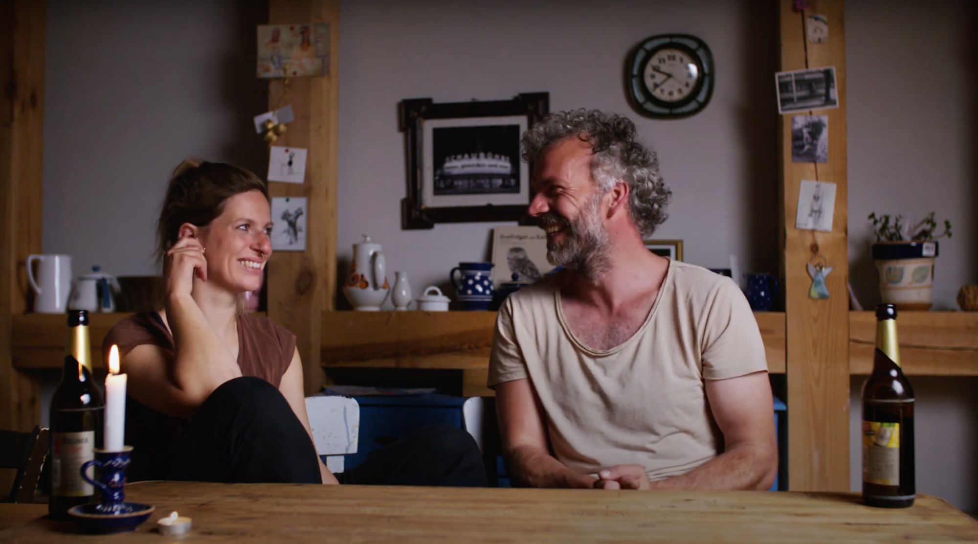Ein Mann und eine Frau lachen sich am Tisch an, im Hintergrund Uhr und Fotos an der Wand