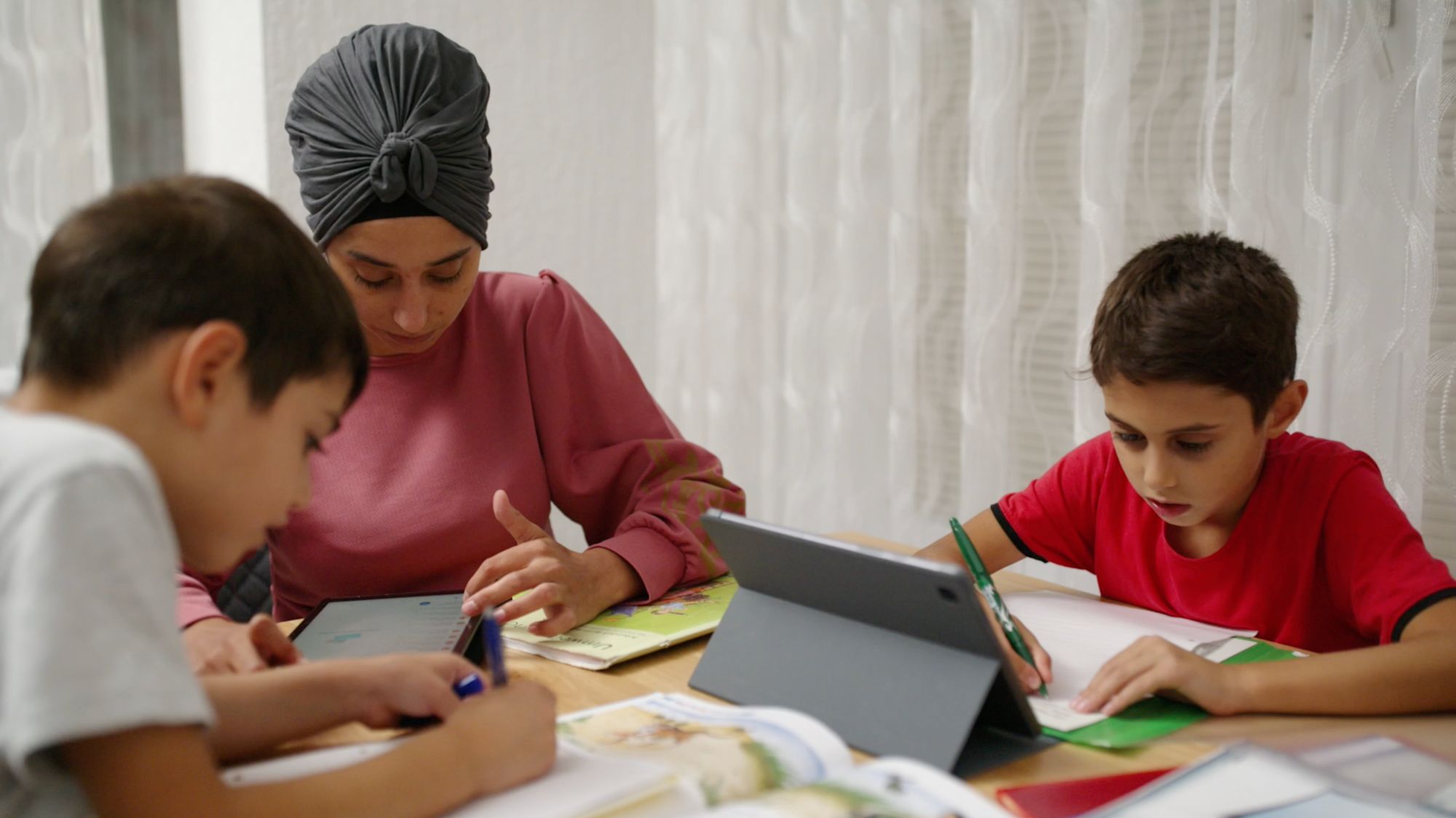 Mutter mit Kopftuch hilft zwei Jungen am Tisch bei den Hausaufgaben, Tablets liegen bereit