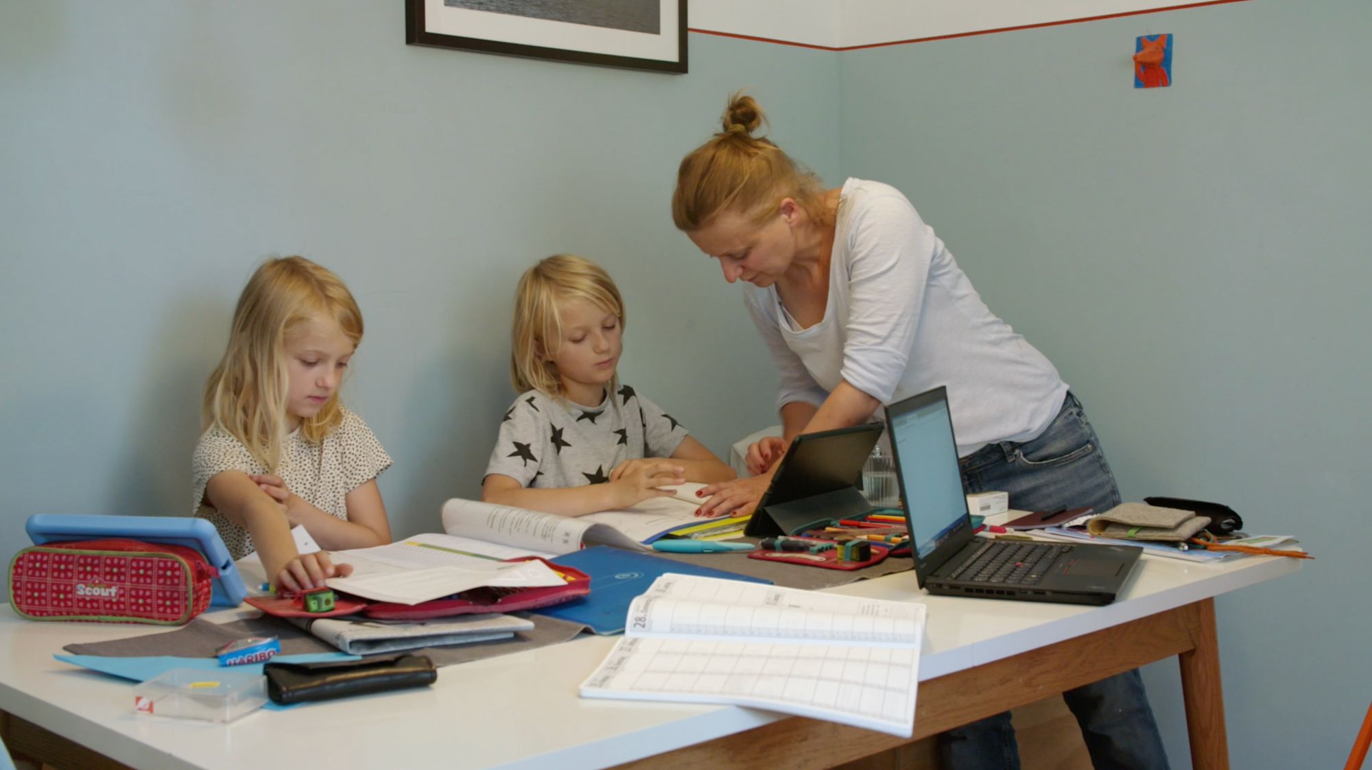 Mutter beugt sich über zwei Kinder am Schreibtisch mit Laptop, Tablet und Schulheften