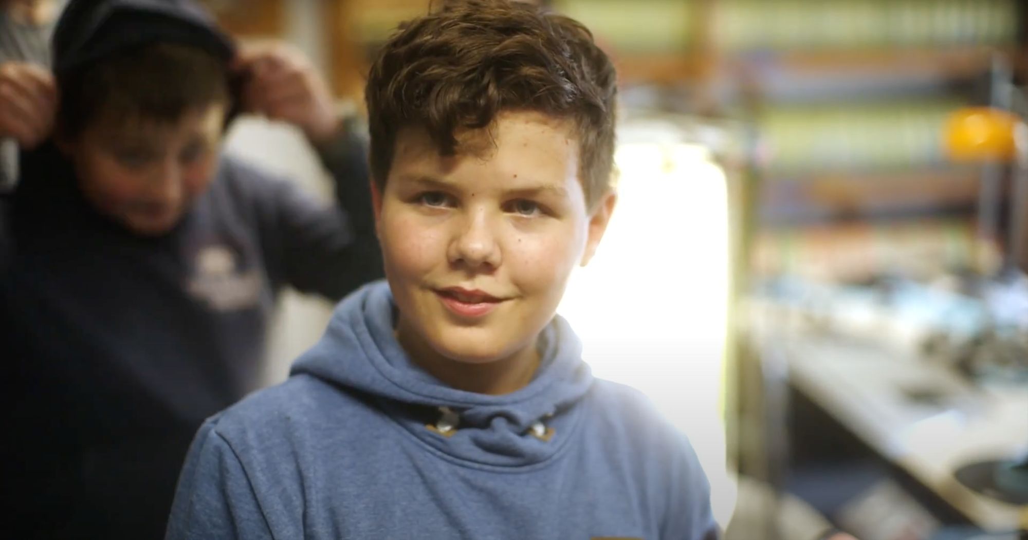 Ein Junge mit lockigem Haar blickt lächelnd in die Kamera, dahinter ein weiterer Junge