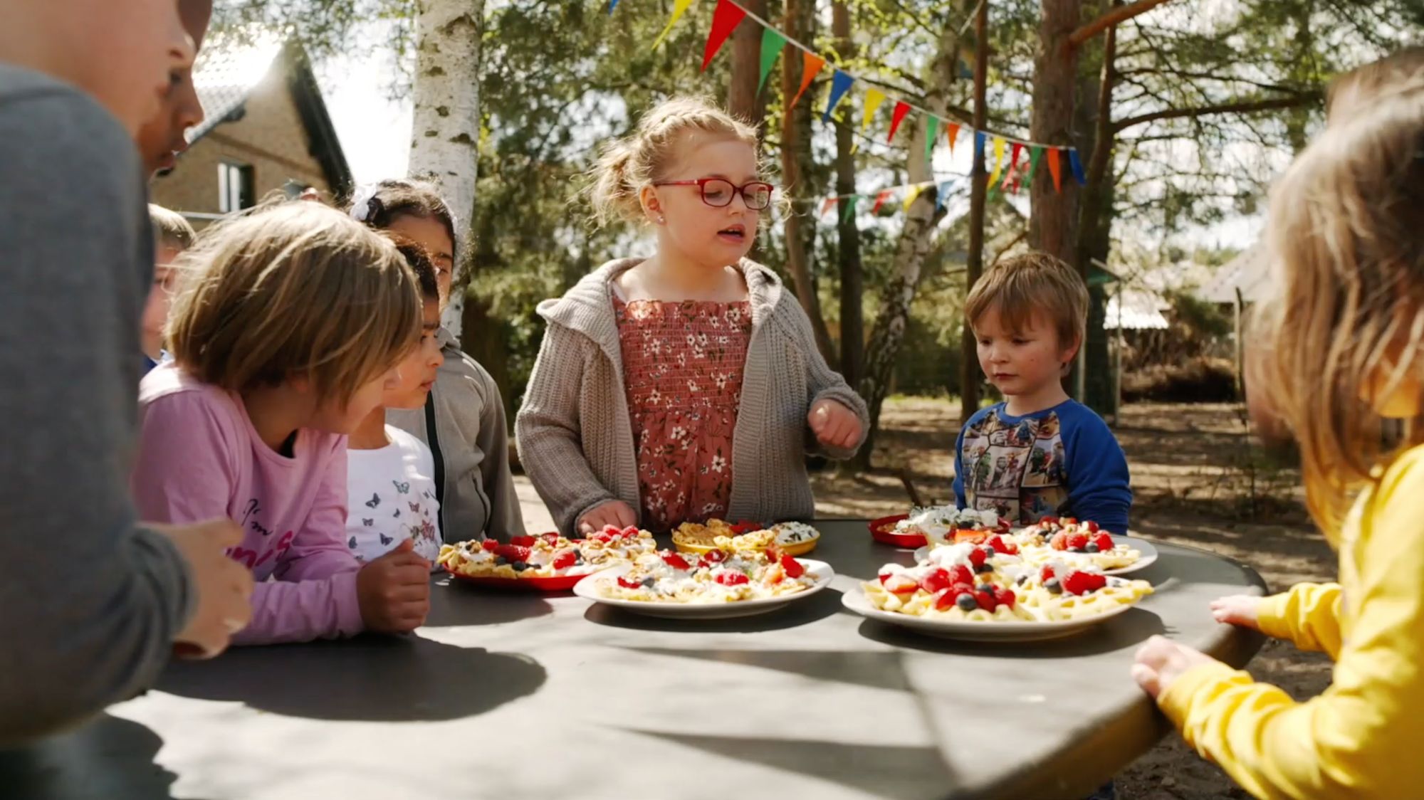 Kinder sitzen an einem Gartentisch mit Waffeln, Beeren und Sahne, Wimpelkette in den Bäumen