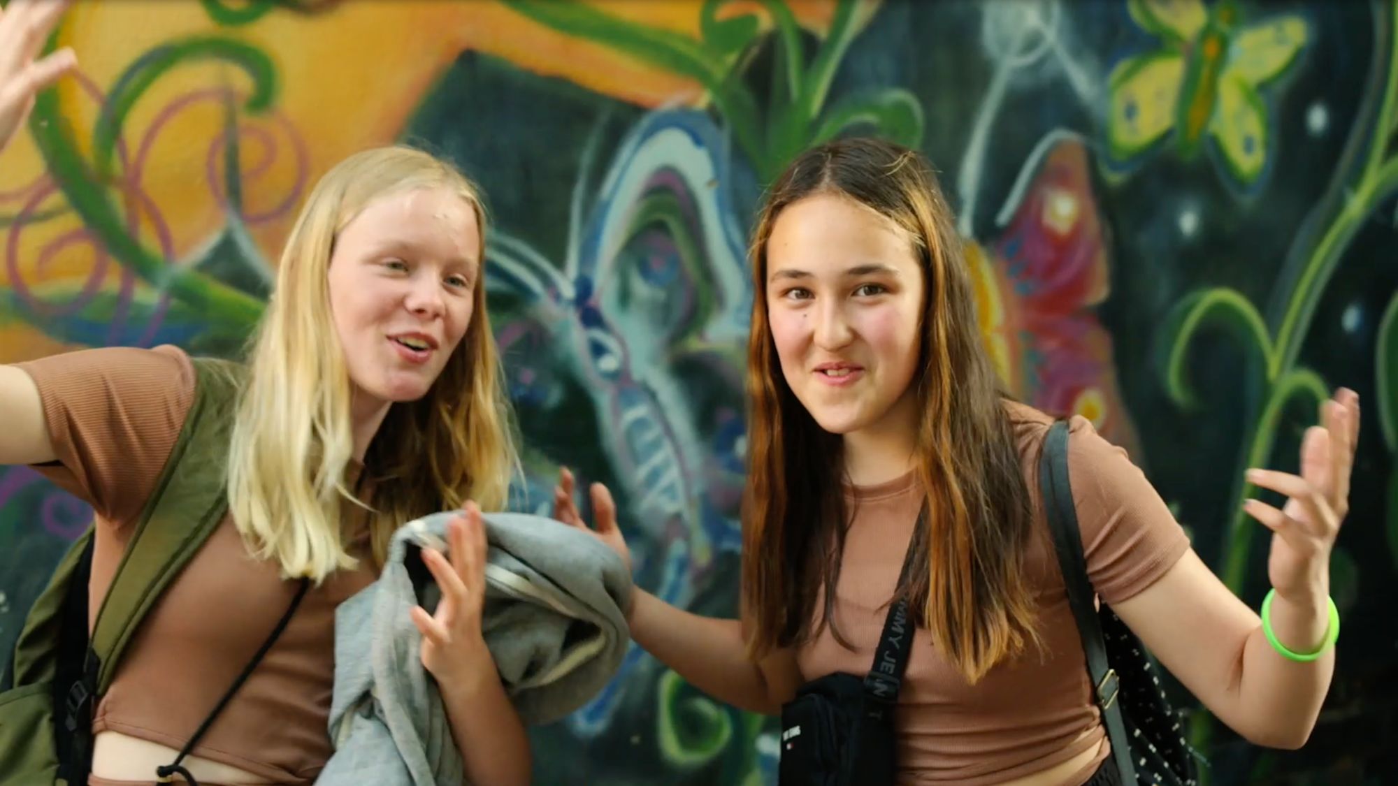 Zwei Mädchen posieren gestikulierend vor einer bunten Graffiti-Wand mit Schmetterlingsmotiv