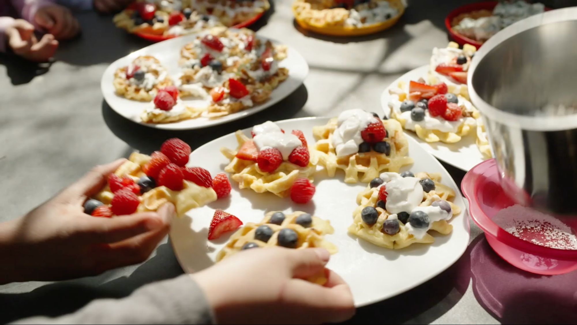 Kinderhände drapieren Erdbeeren und Blaubeeren auf Waffeln mit Sahnehäubchen