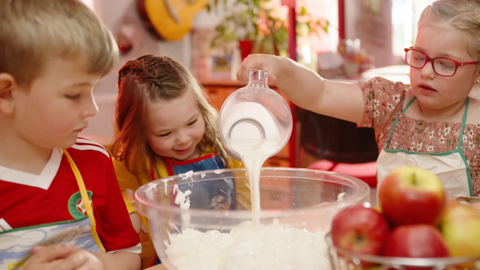 Drei Kinder in Schürzen gießen Sahne über geraspelte Kokosflocken in einer Glasschüssel