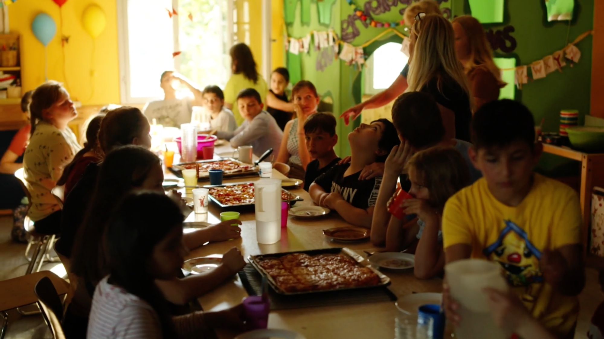 Große Kindergruppe sitzt an einer langen Tafel mit Pizza in bunt dekoriertem Raum