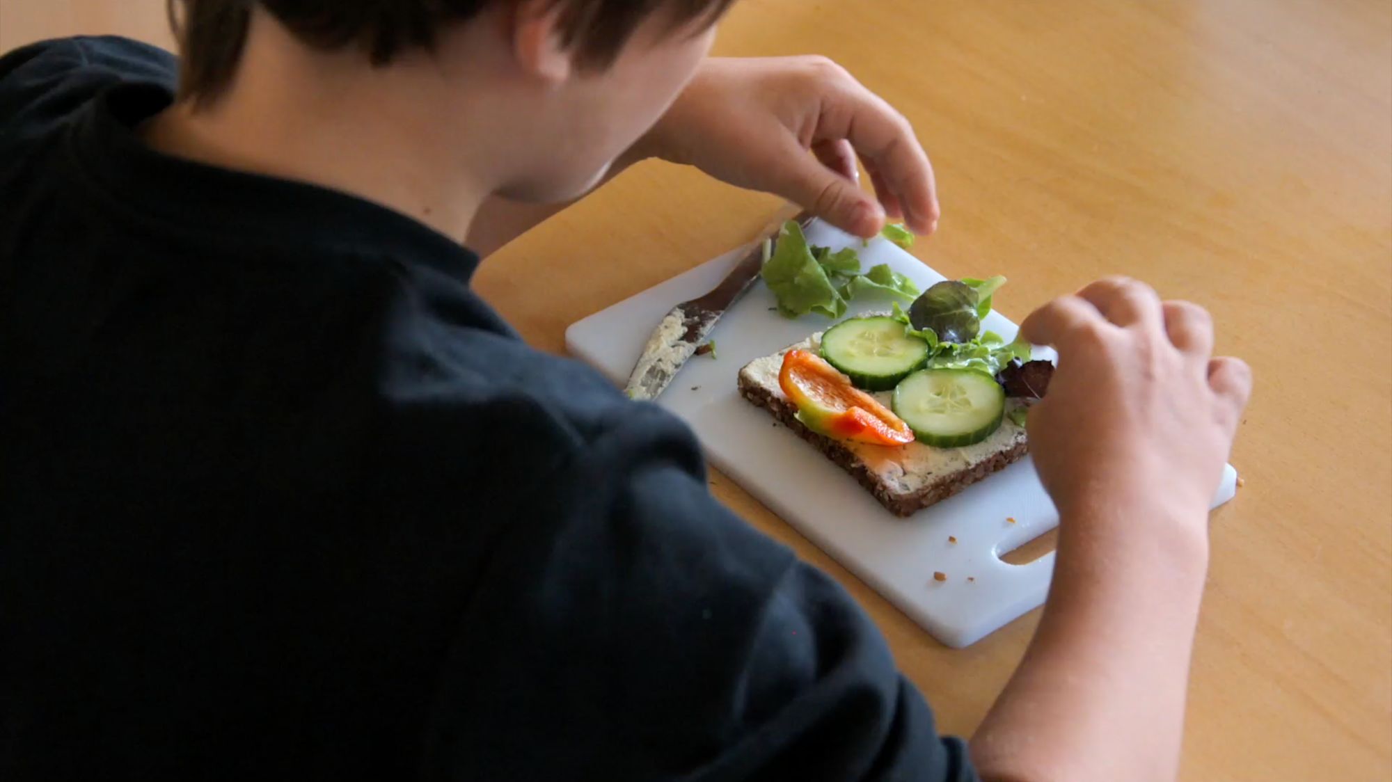 Kind belegt von oben fotografiert ein Vollkornbrot mit Gurke, Paprika und Salat