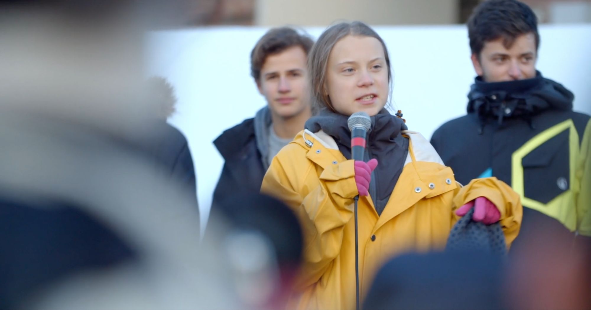 Junge Frau in gelber Jacke spricht am Mikrofon vor einer Gruppe Jugendlicher