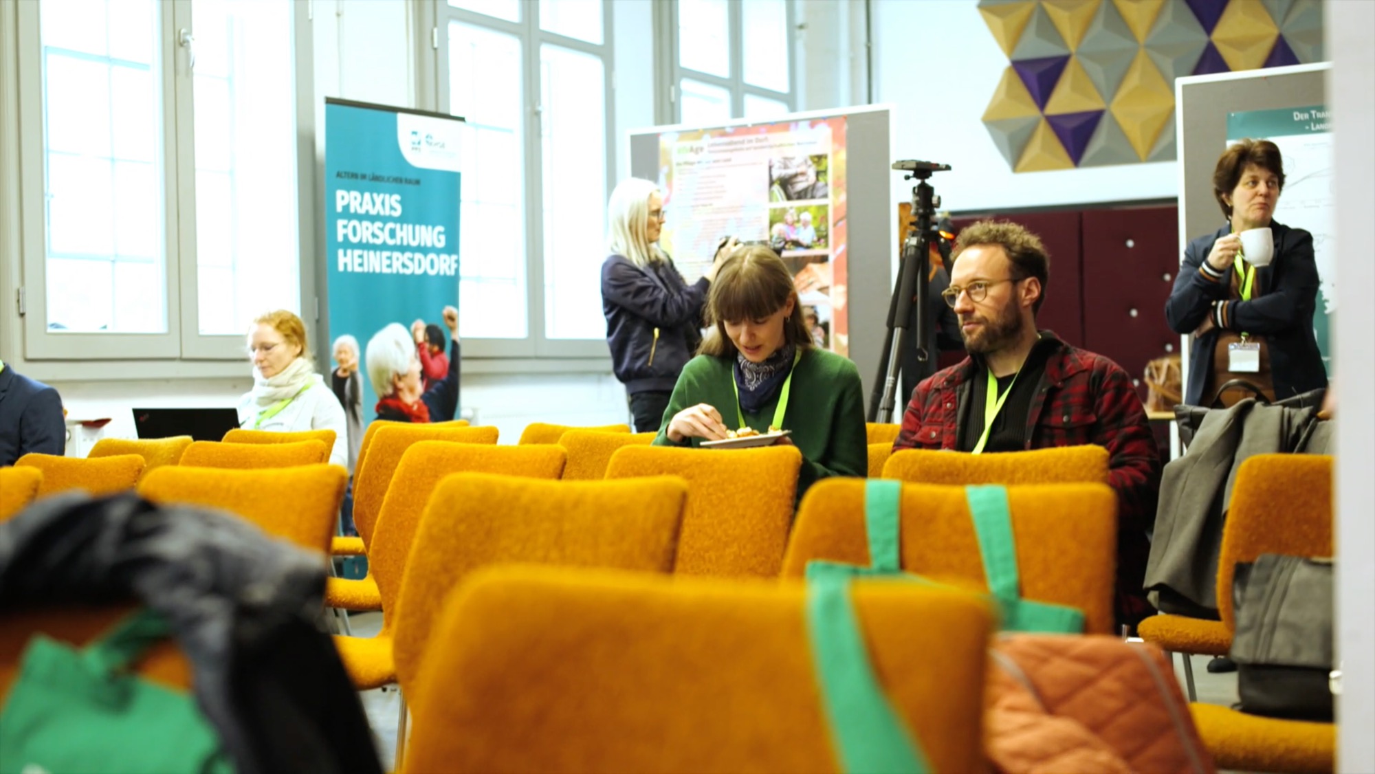 Participants sit in an event room with banners for "Praxisforschung Heinersdorf"