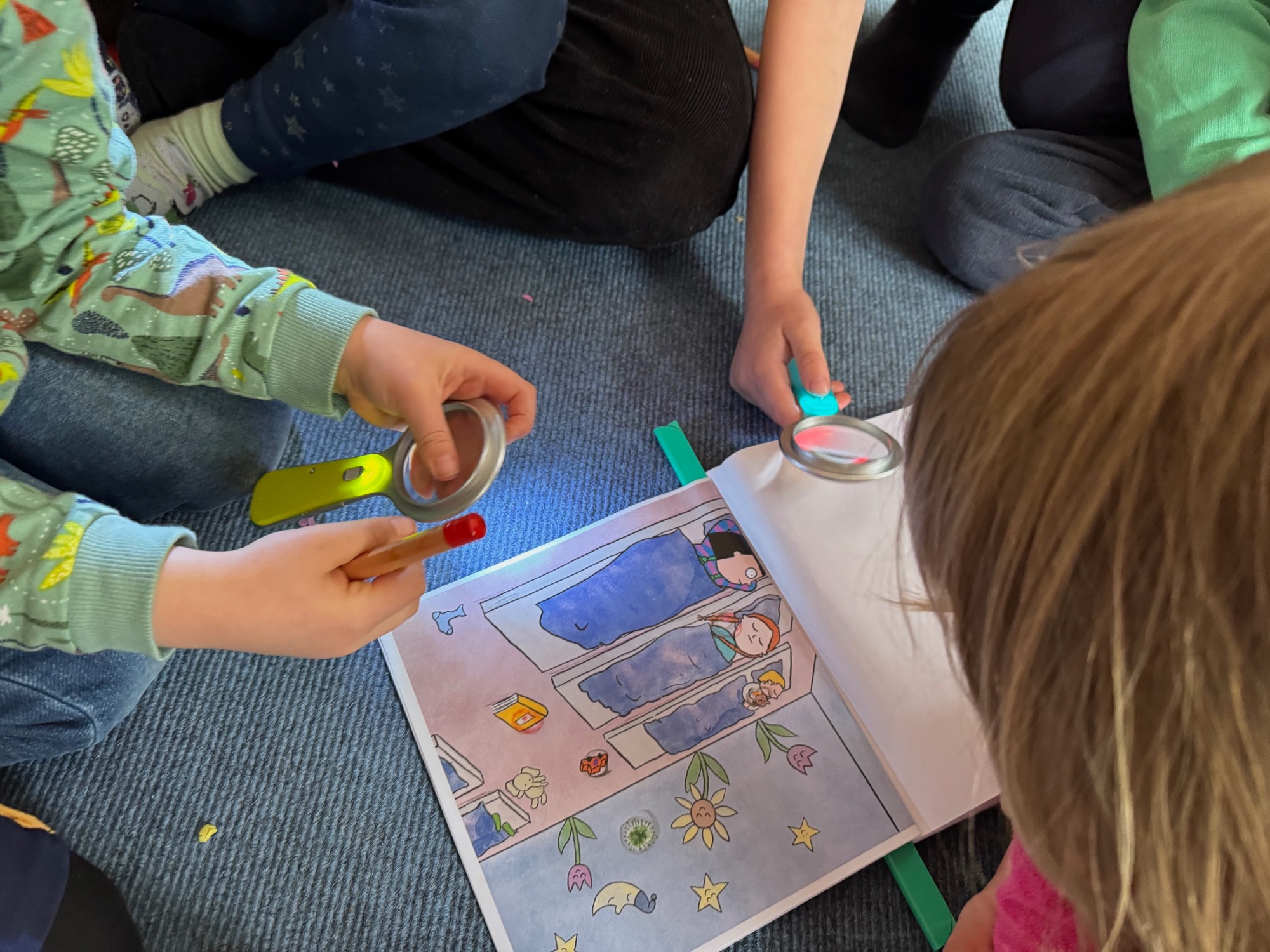 Zwei Kinder betrachten mit Lupen eine illustrierte Buchseite mit schlafenden Figuren