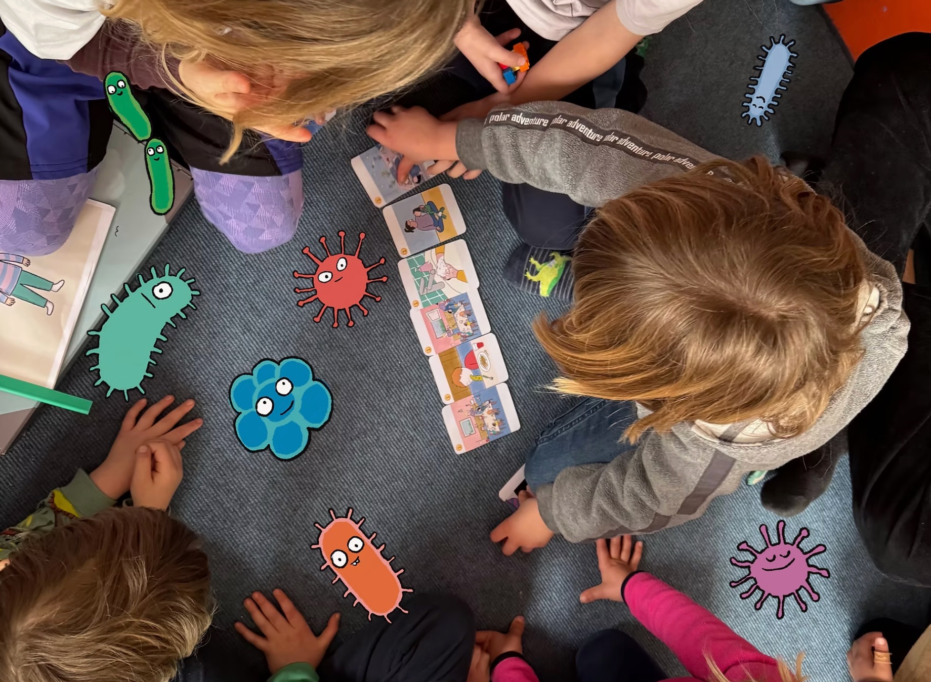 Luftaufnahme einer Kindergruppe im Kreis um Bildkarten, umringt von aufgeklebten Mikroben-Illustrationen