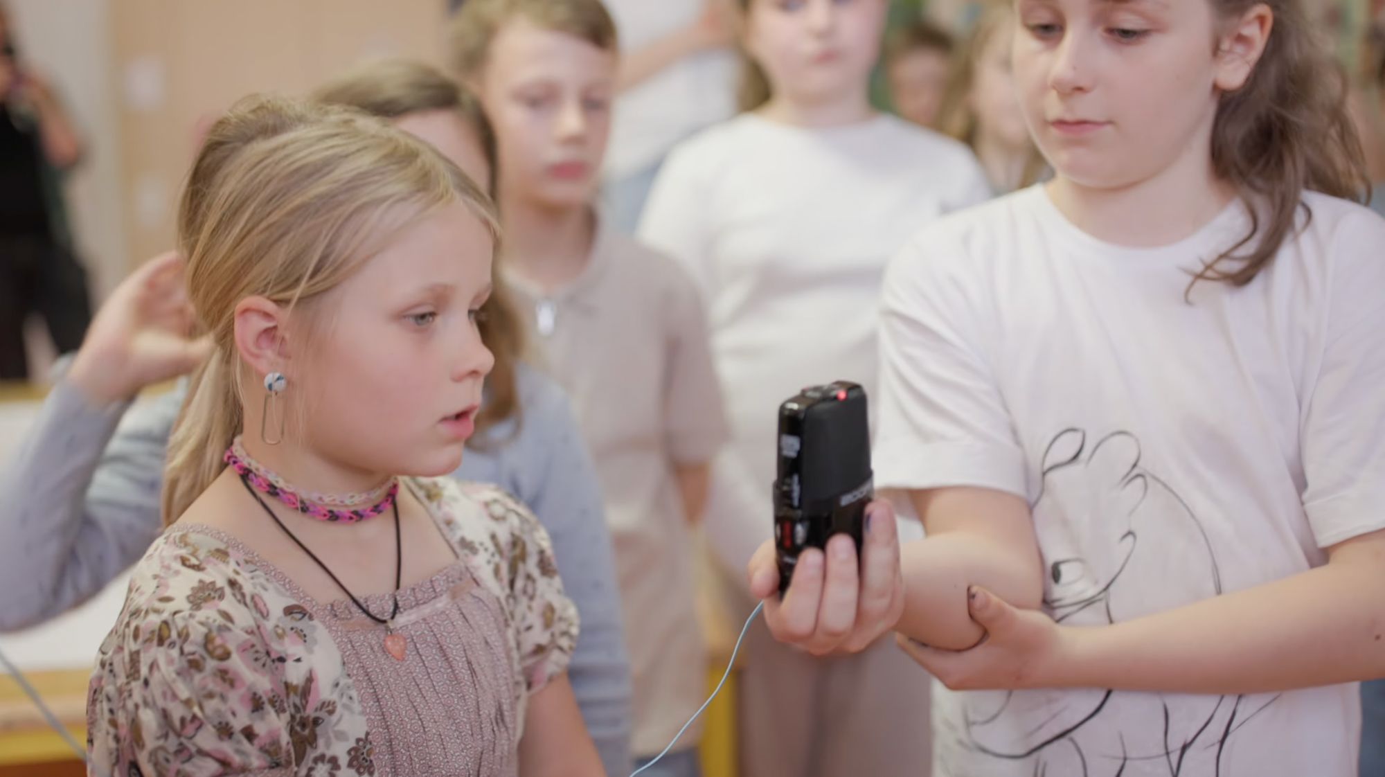 Mädchen mit Kette hält ein Aufnahmegerät, während sie spricht, Kinder im Hintergrund