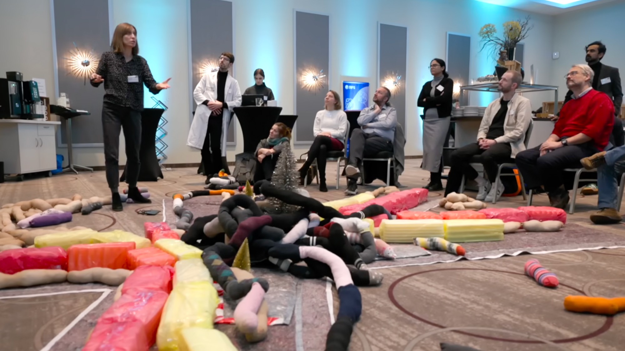 Workshop-Teilnehmende sitzen im Kreis um eine bunte Boden-Installation aus gepolsterten, schlangenförmigen Objekten
